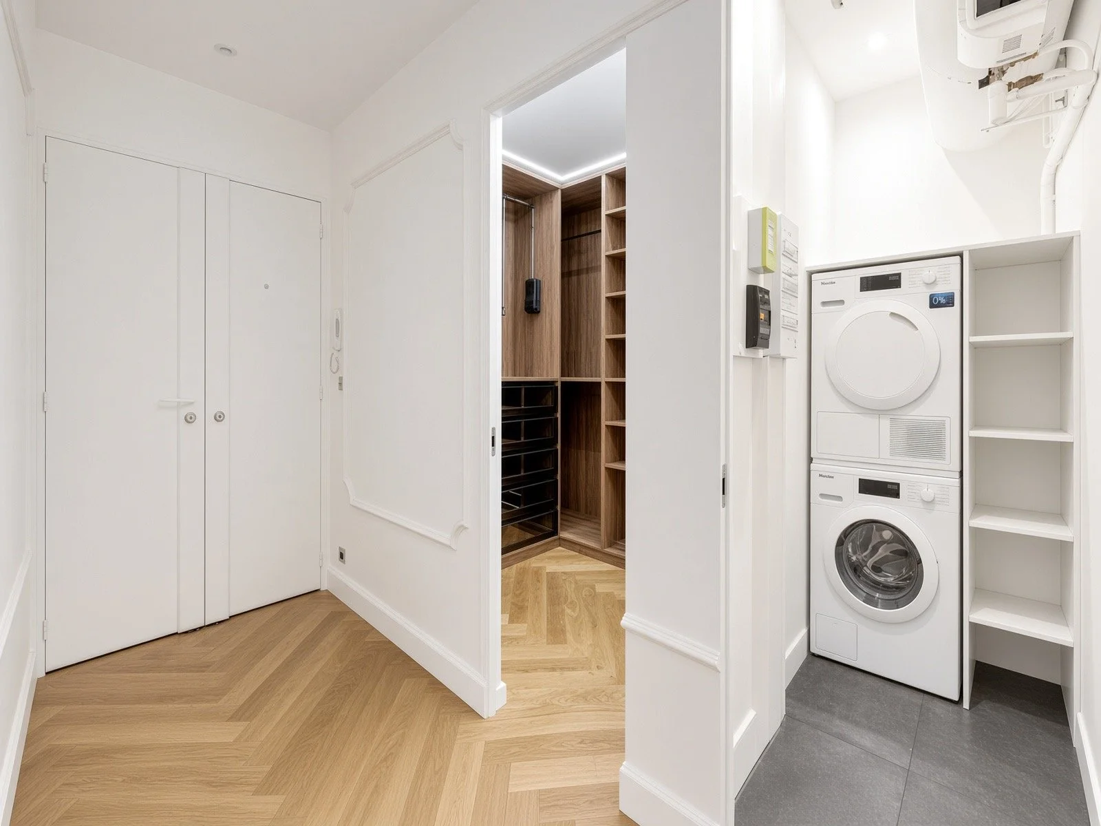 A laundry room with stacked washer and dryer, white shelving unit, textured gray tile floors, and white walls.