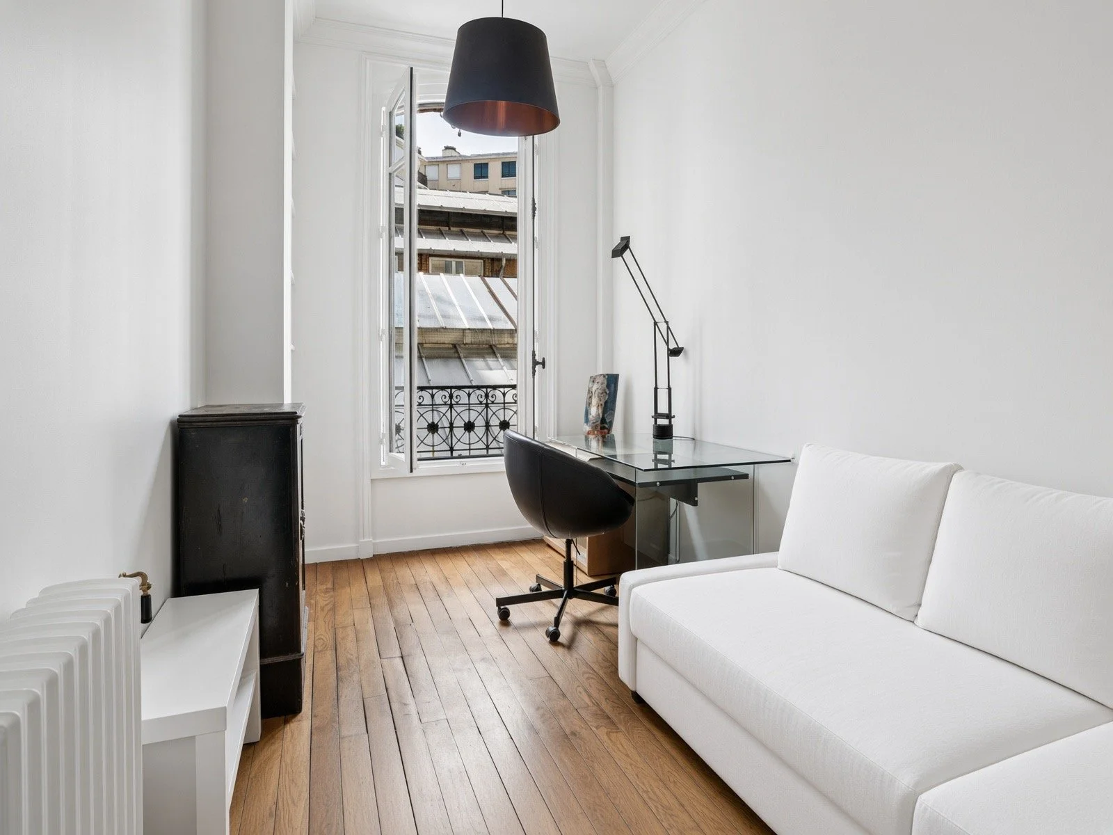 A small, minimalist home office with a white sofa, a glass desk, and a black swivel chair. Open window with a view of rooftops and a black pendant light hanging from the ceiling.