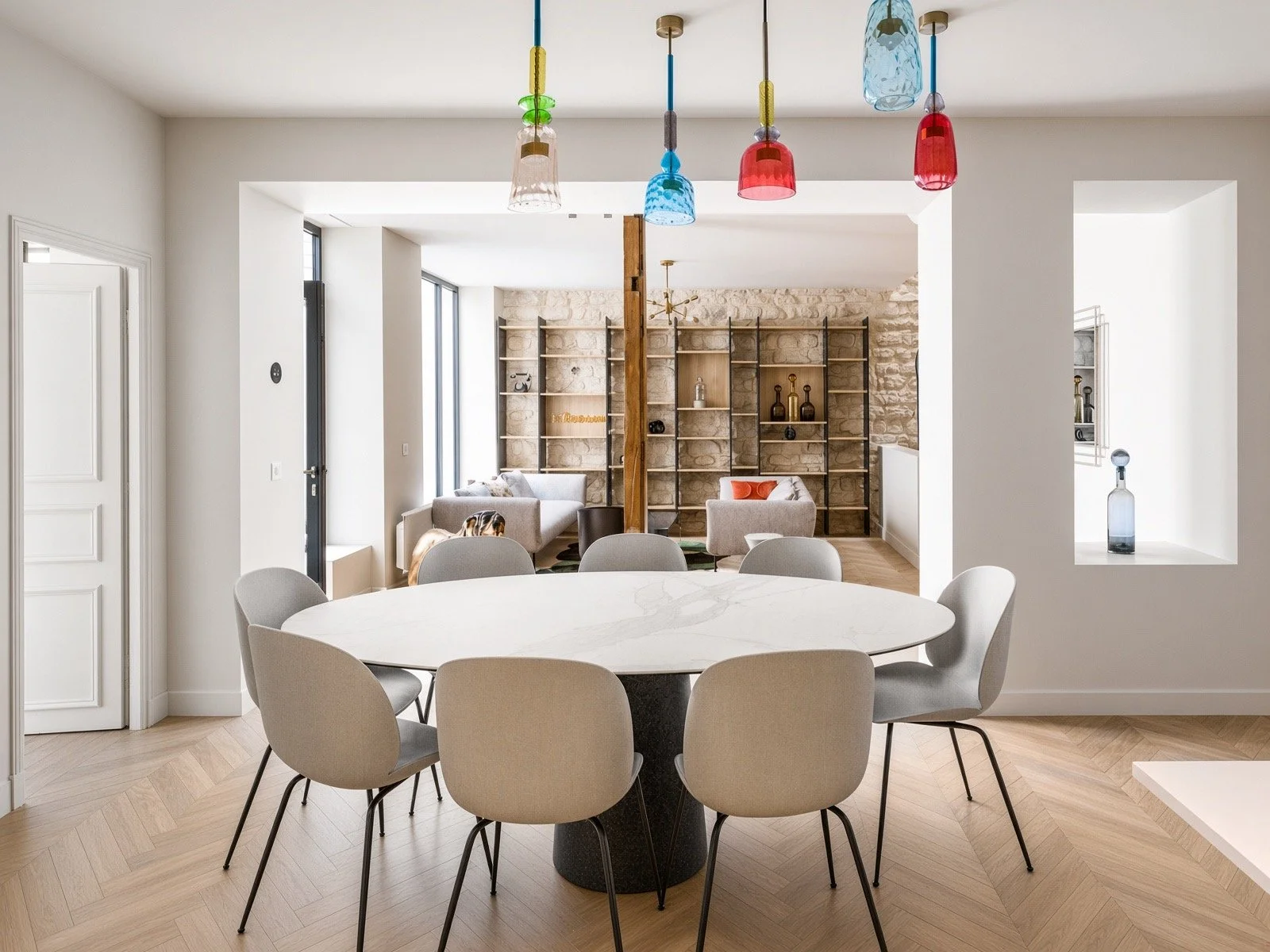 Modern dining room with a white round table and beige chairs, colorful hanging light fixtures, and a living area with sofas and shelves in the background.