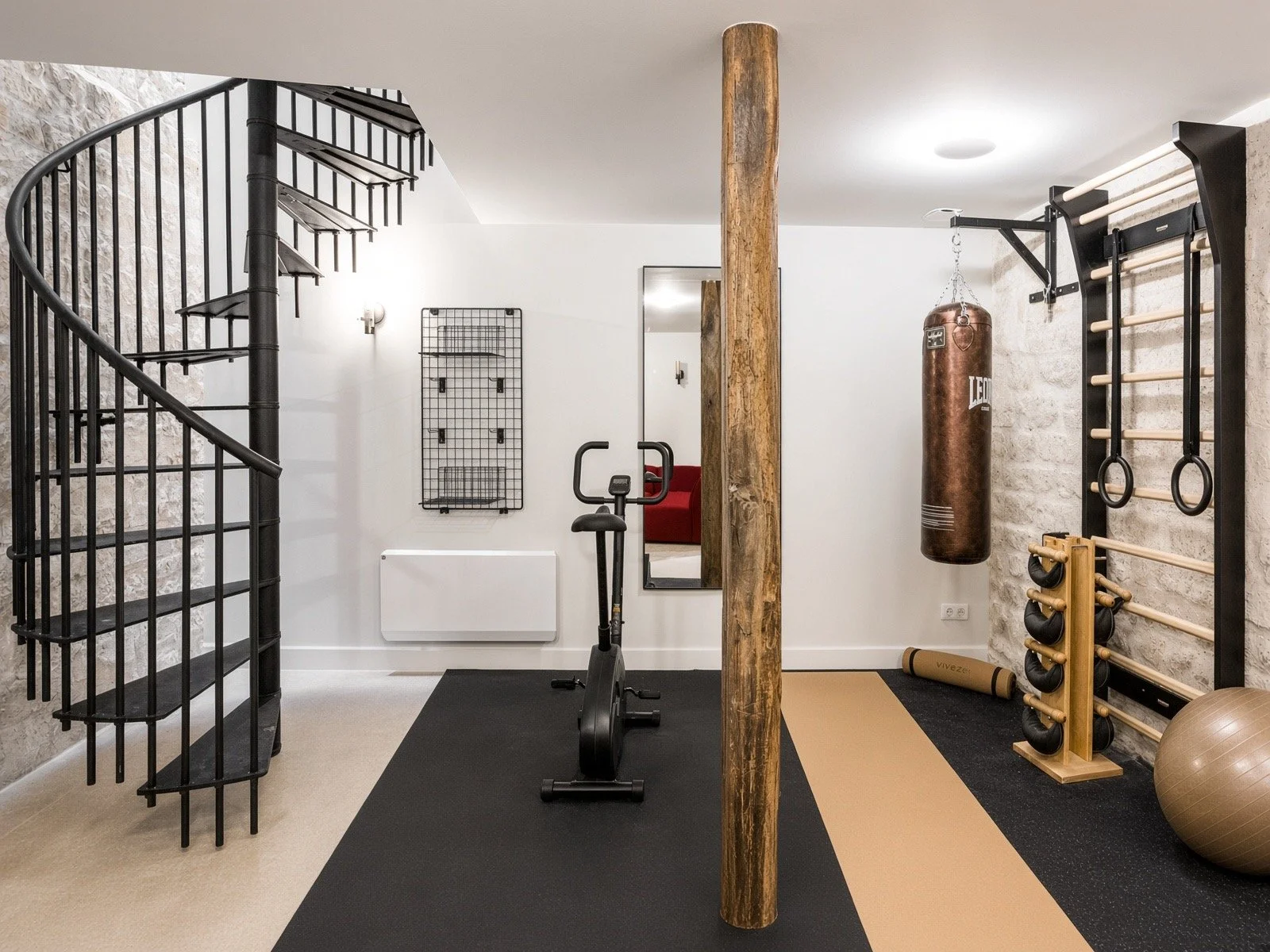 Home gym room with boxing punching bag, exercise equipment, wooden wall bar, fitness weights, and a spiral staircase, with a white wall and stone wall decor.