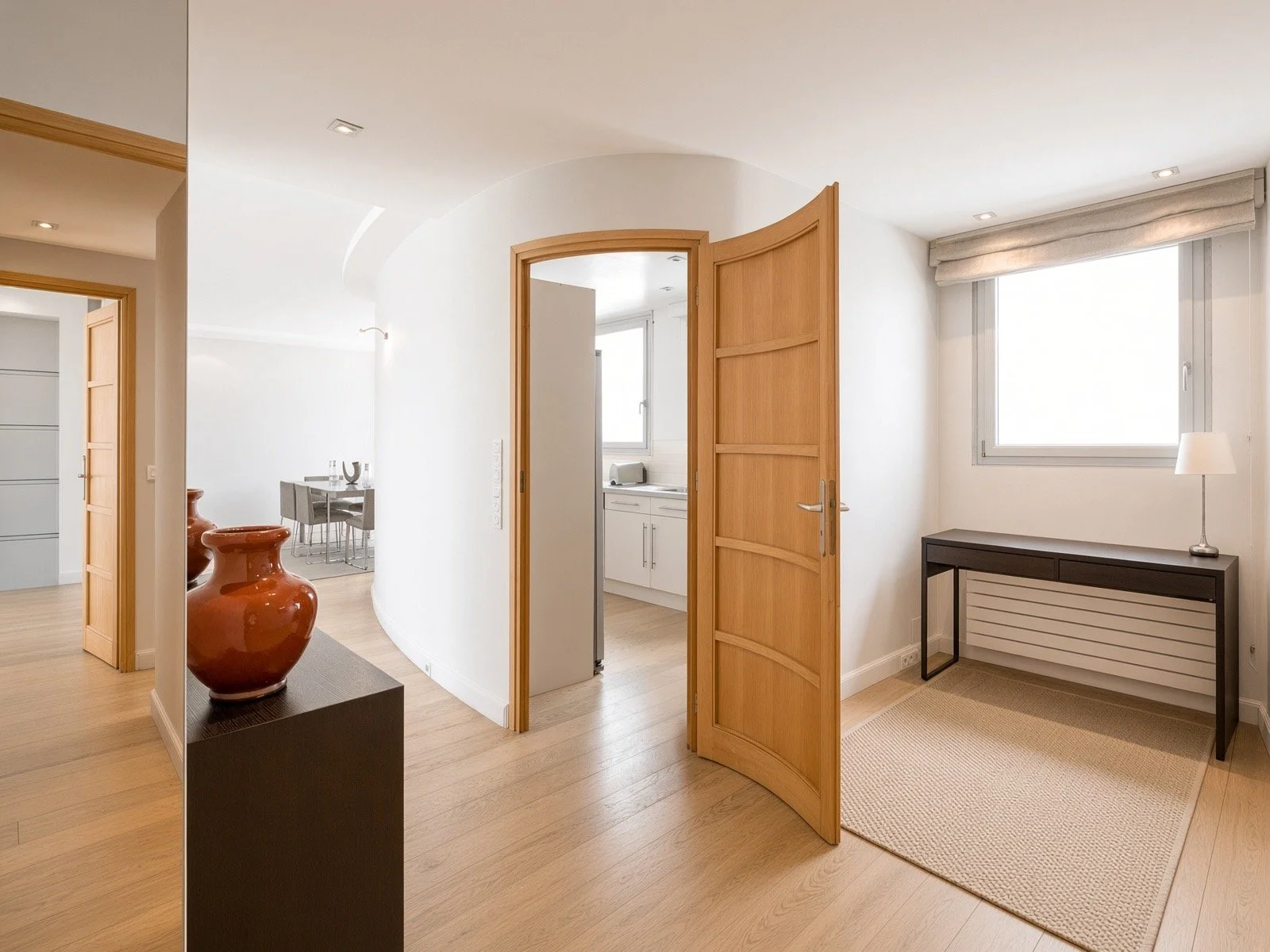 Interior view of a modern apartment with wooden flooring, white walls, open doors leading to a kitchen and dining area, and decorative vases on a black pedestal in the foreground.