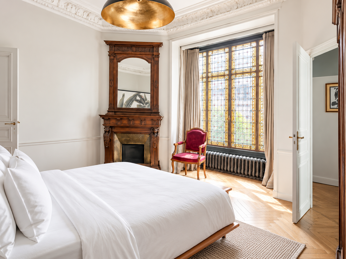 A bedroom with a large bed covered in white bedding, a vintage wooden mirror above a fireplace, a red velvet armchair near a window with stained glass, beige curtains, a radiator, and hardwood floors.