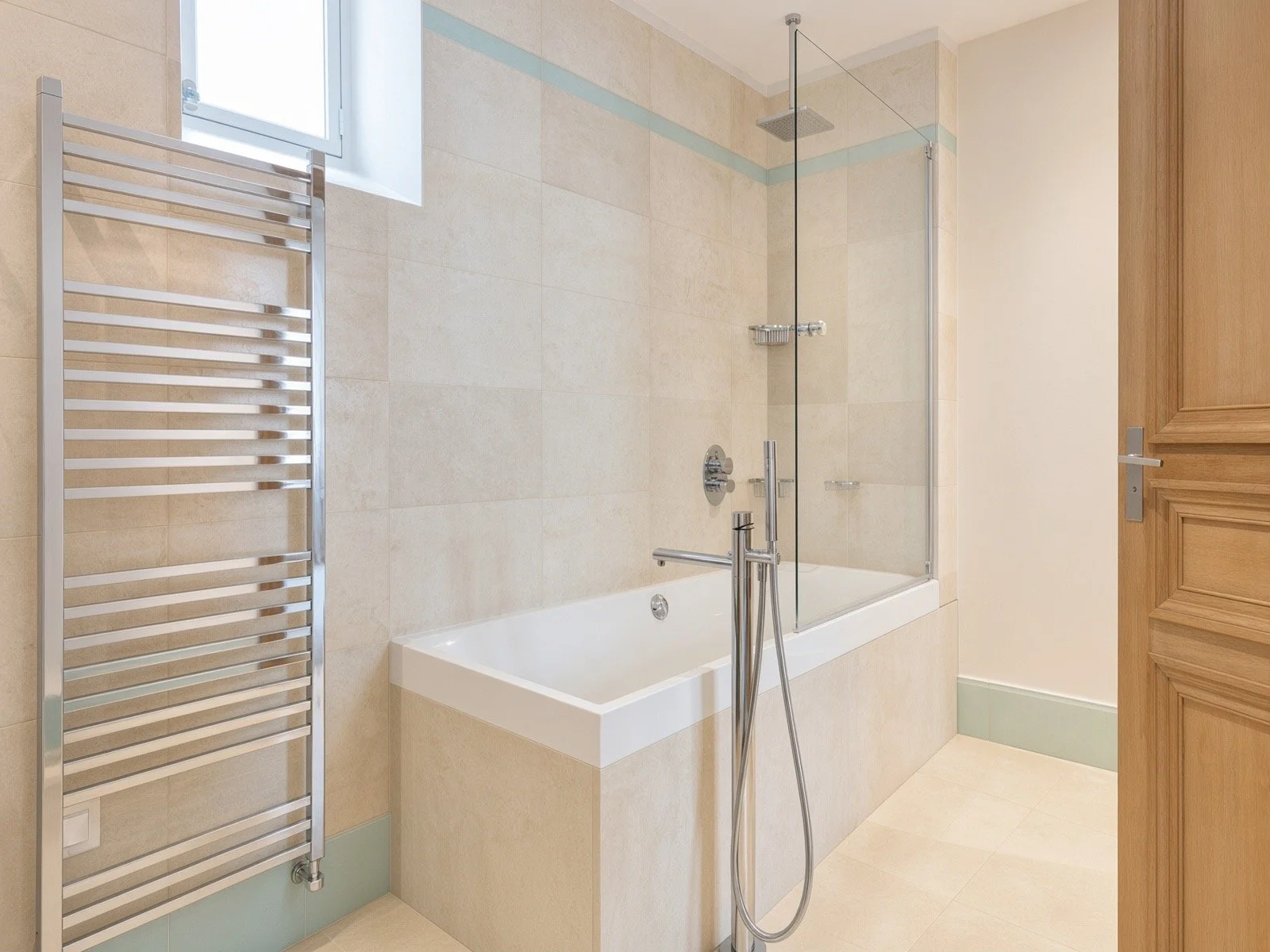 A modern bathroom with a bathtub, showerhead, glass shower partition, a window, a heated towel rack, beige tiles, and a wooden door.