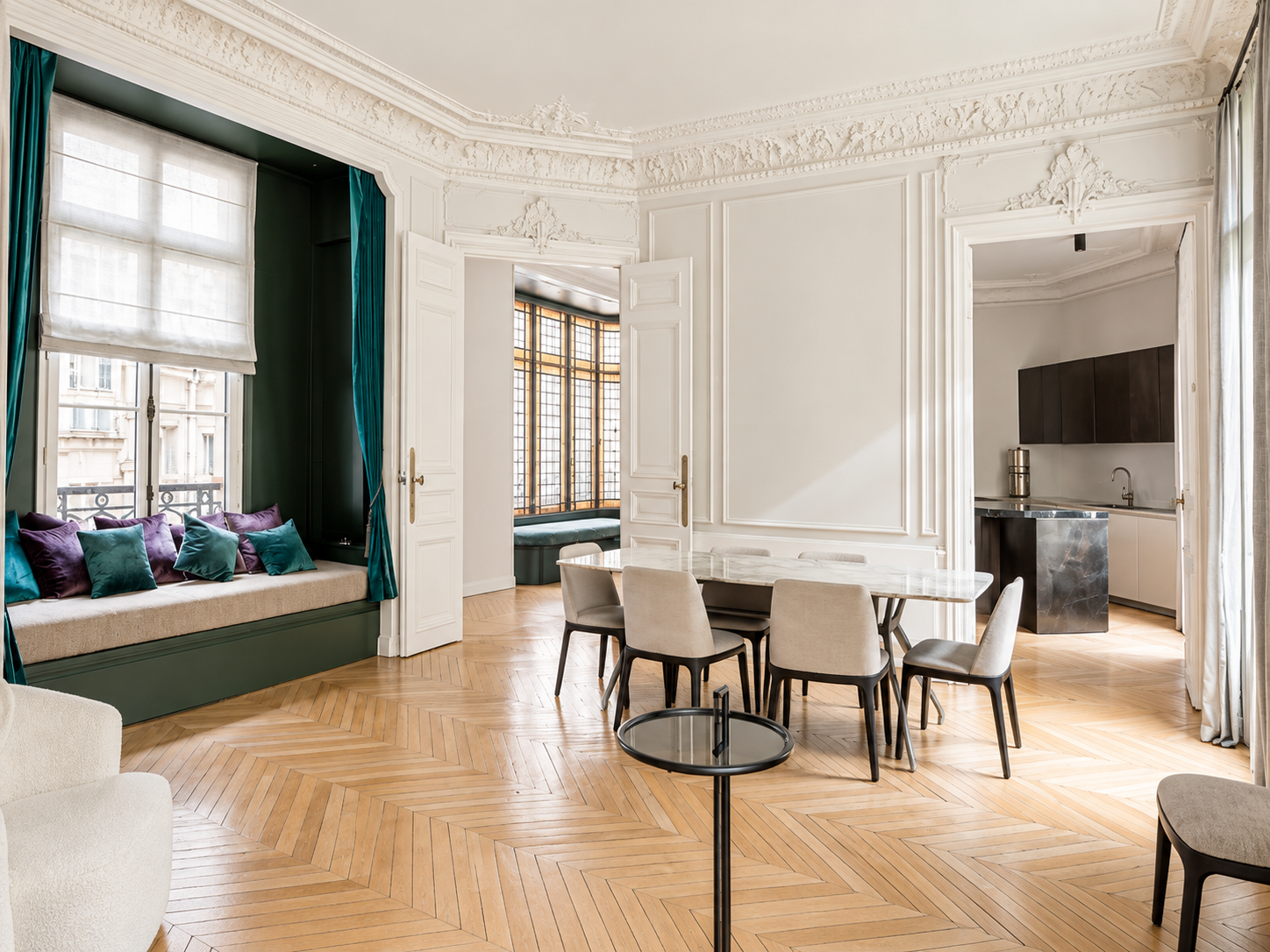 Elegant living space with wooden herringbone floors, built-in window seat with purple and teal cushions, white walls with decorative molding, large windows with curtains, a dining table with beige chairs, and a small kitchen area in the background.
