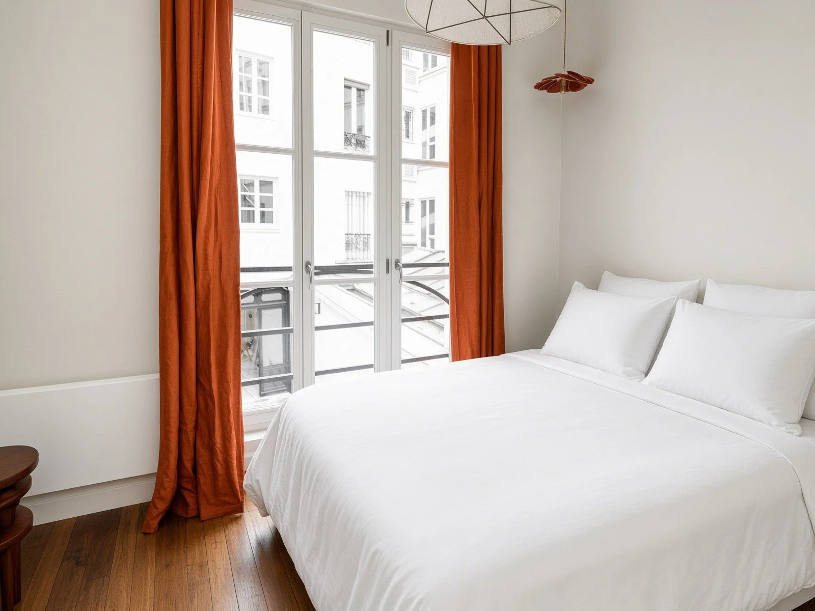 A bright bedroom with a large bed covered in white linens, orange curtains, and a view of adjacent white buildings through a glass door.