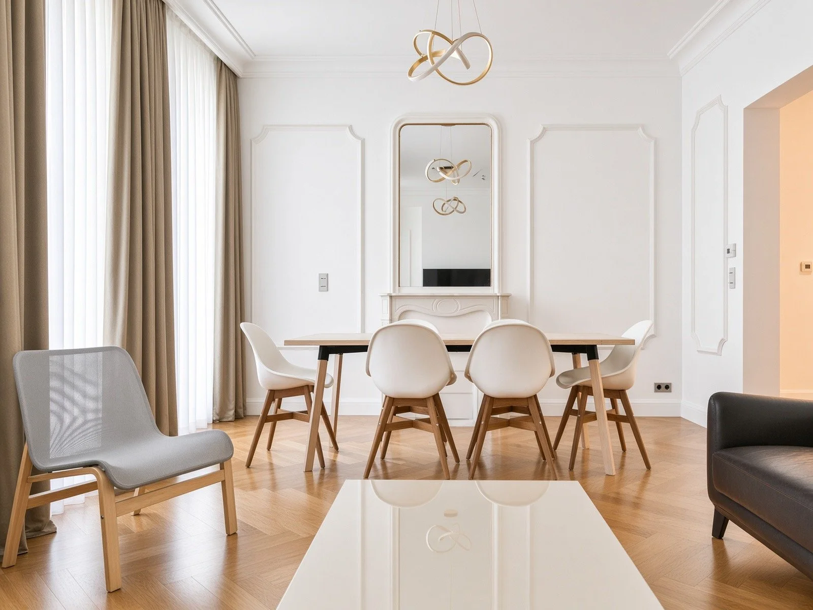 Modern dining room with wooden floor, white walls, and beige curtains. A rectangular wooden dining table with six white chairs. Minimalist lighting fixture hanging from the ceiling. A mirror above a fireplace mantel on the wall.