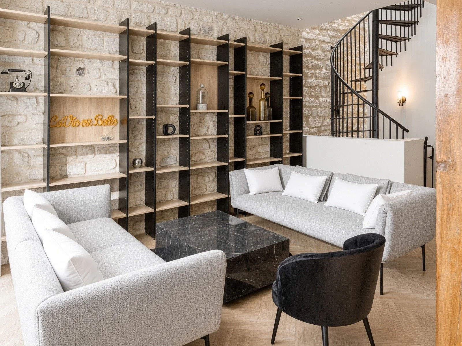 Modern living room with gray sofas, decorative black armchair, black marble coffee table, and tall bookshelf with decorative items against a stone wall.
