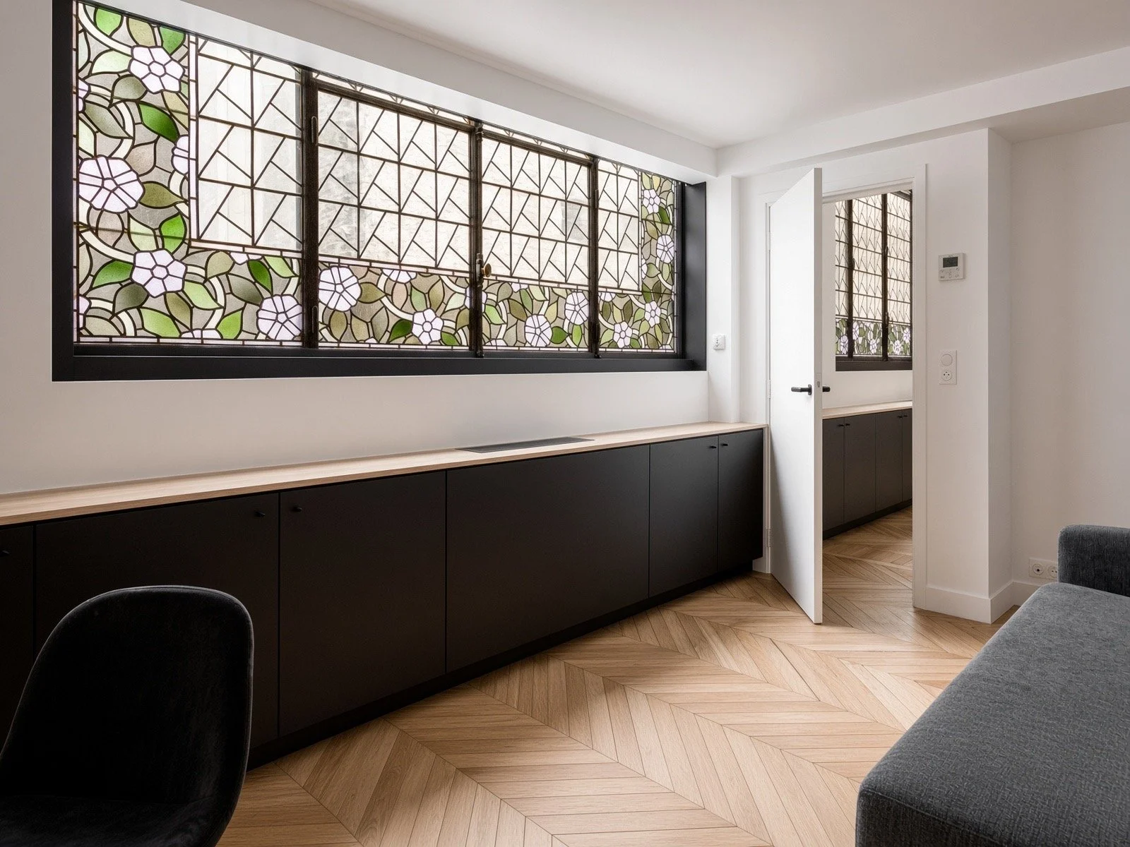 Modern interior room with large stained glass window featuring floral design, black cabinetry, wooden flooring, and a doorway leading to another room.