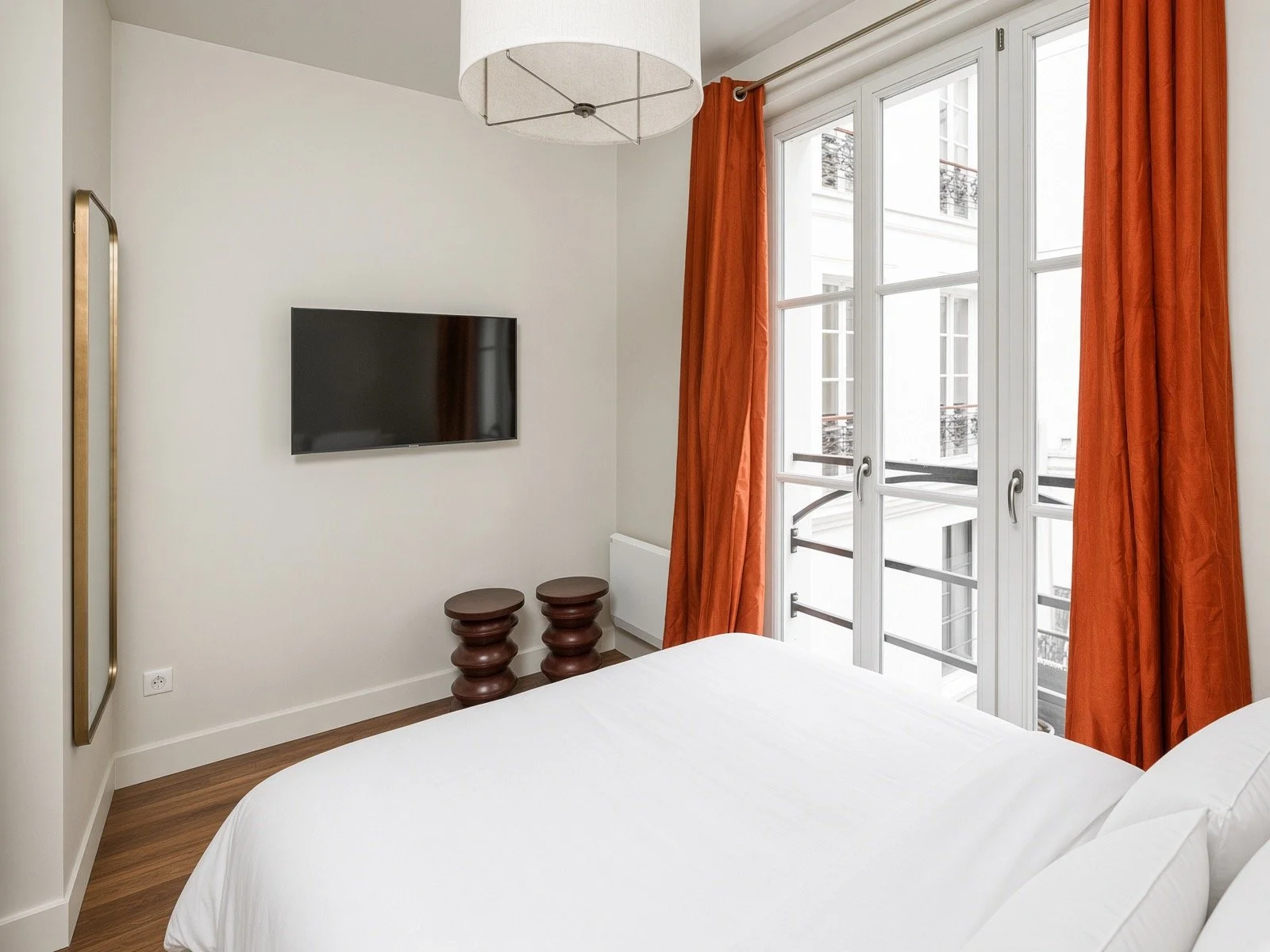 A bedroom with a large window with orange curtains, a wall-mounted TV, two small wooden stools, and a white bed.