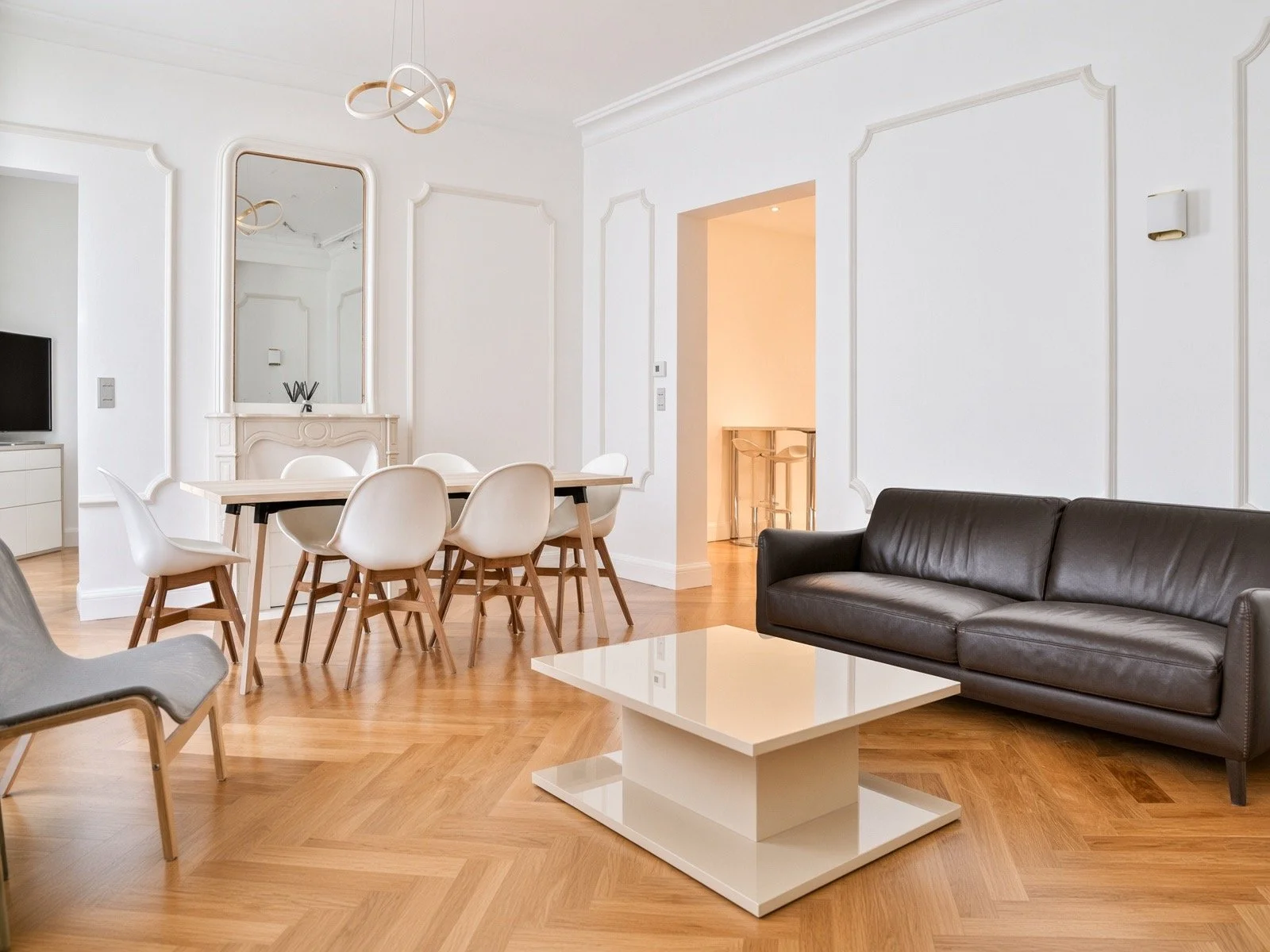 Modern living room with white walls, wooden flooring, a black leather sofa, a white coffee table, a dining table with six white chairs, a large mirror, and a ceiling light fixture.