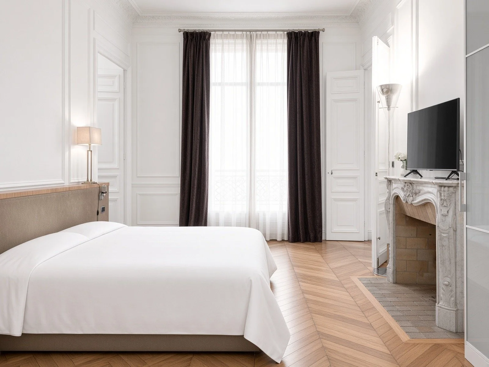 Elegantly decorated hotel room with white walls and ornate molding, featuring a large window with sheer curtains and dark drapes, a bed with white bedding, a modern bedside lamp, a flat-screen TV on a white decorative fireplace mantel, and hardwood flooring.