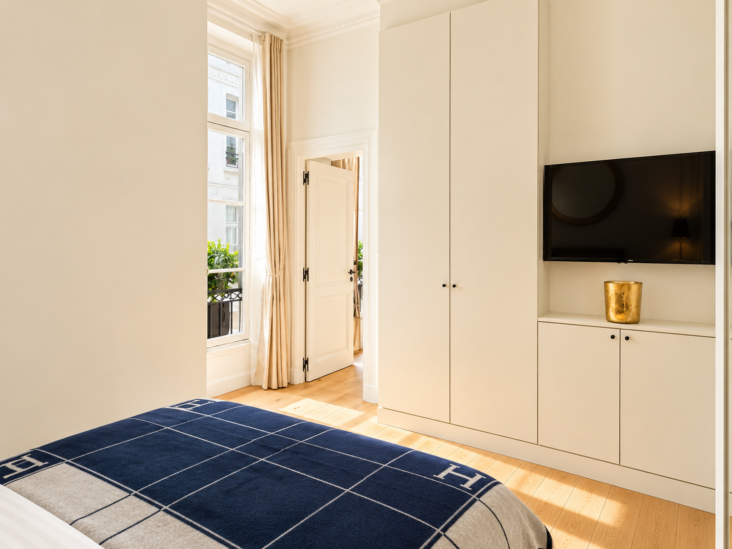 A well-lit bedroom corner with a bed partially visible in the foreground, featuring a blue and white checkered blanket. To the right, built-in white cabinetry with a mounted flat-screen TV and a gold decorative pot. Behind the bed, a window with cream curtains and a door leading to a balcony or outside area with potted plants is visible. Light wood flooring completes the scene.