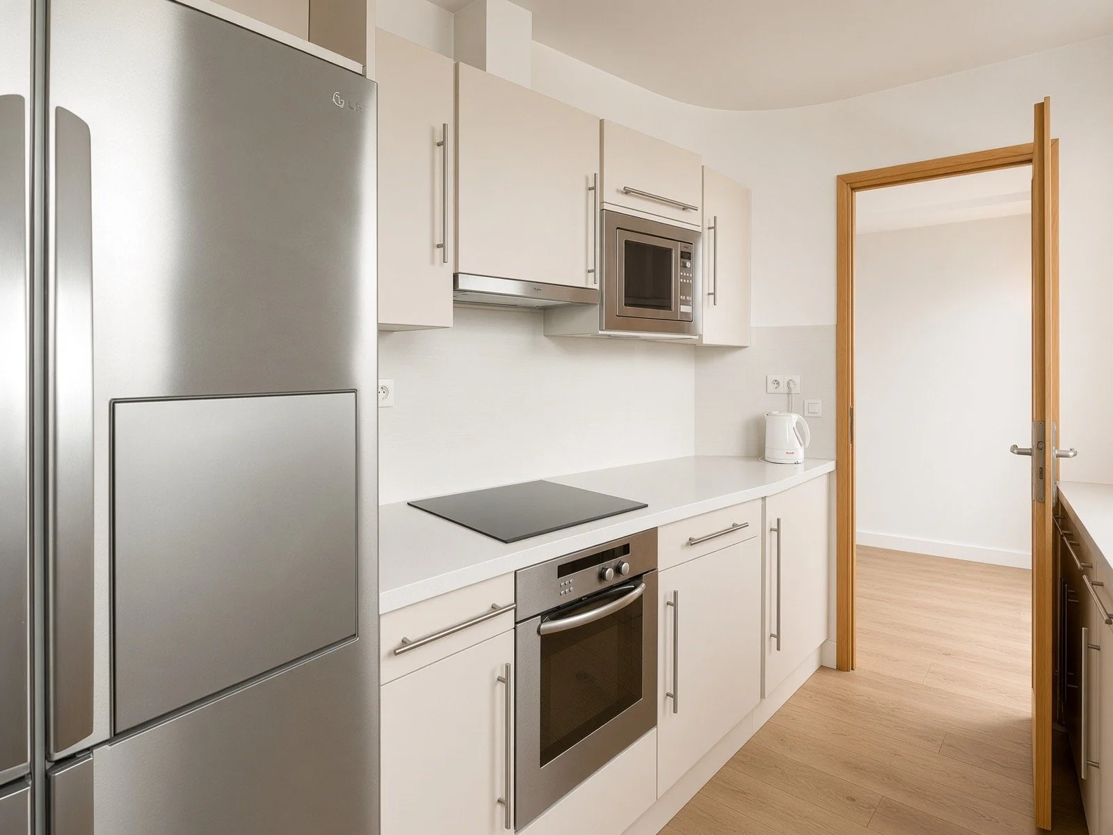 Modern kitchen with white cabinets, stainless steel appliances including fridge, oven, microwave, and electric cooktop, a white countertop, and a kettle. There's a doorway leading to another room with light-colored flooring.
