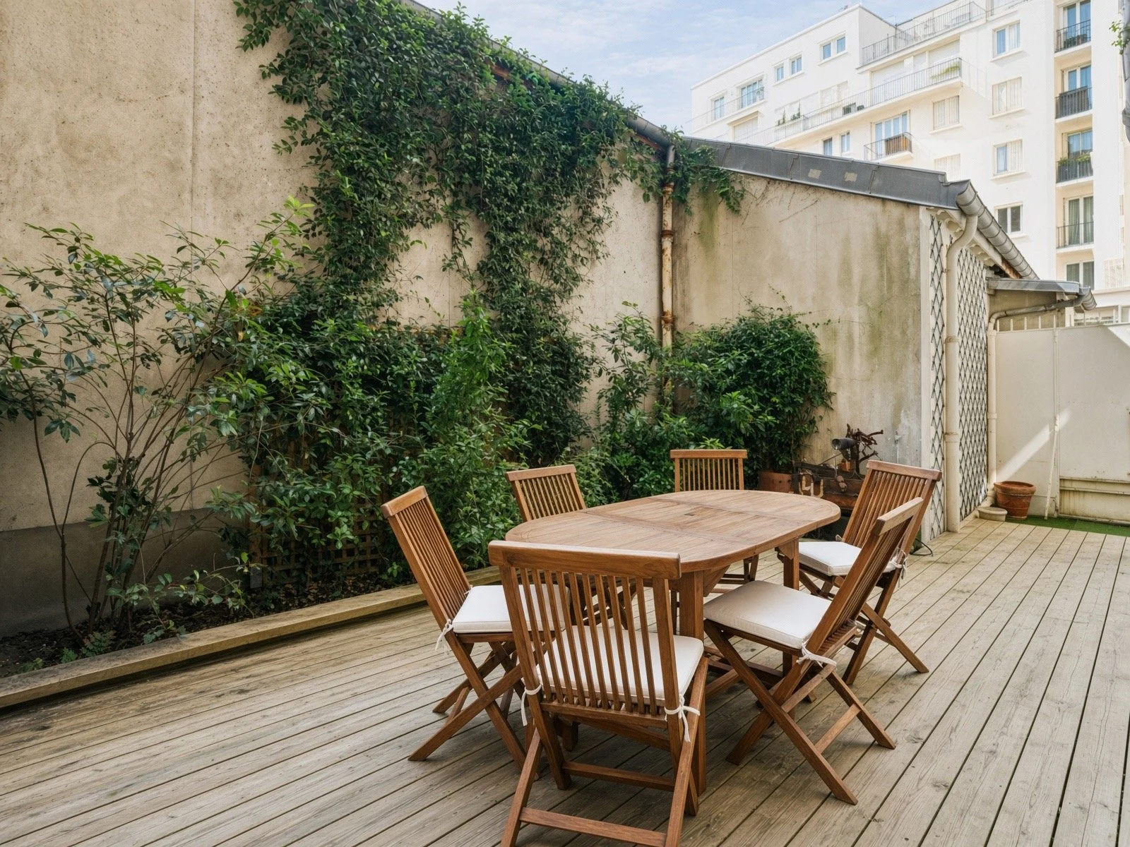 Outdoor patio with wooden table and chairs, surrounded by plants and greenery, in an urban setting with nearby apartment buildings.