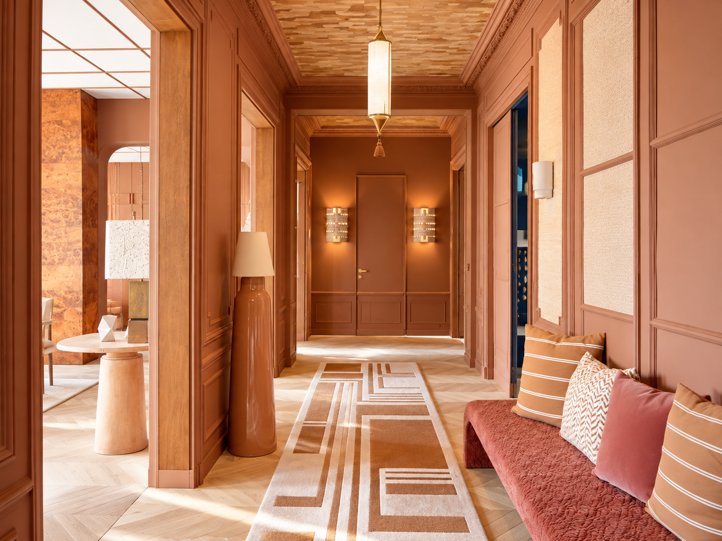 Elegant hotel hallway with wood paneling, a patterned rug, and modern lighting. A sitting area with pillows and a long cushion is visible on the right, and a side table with a tall ceramic lamp is on the left.