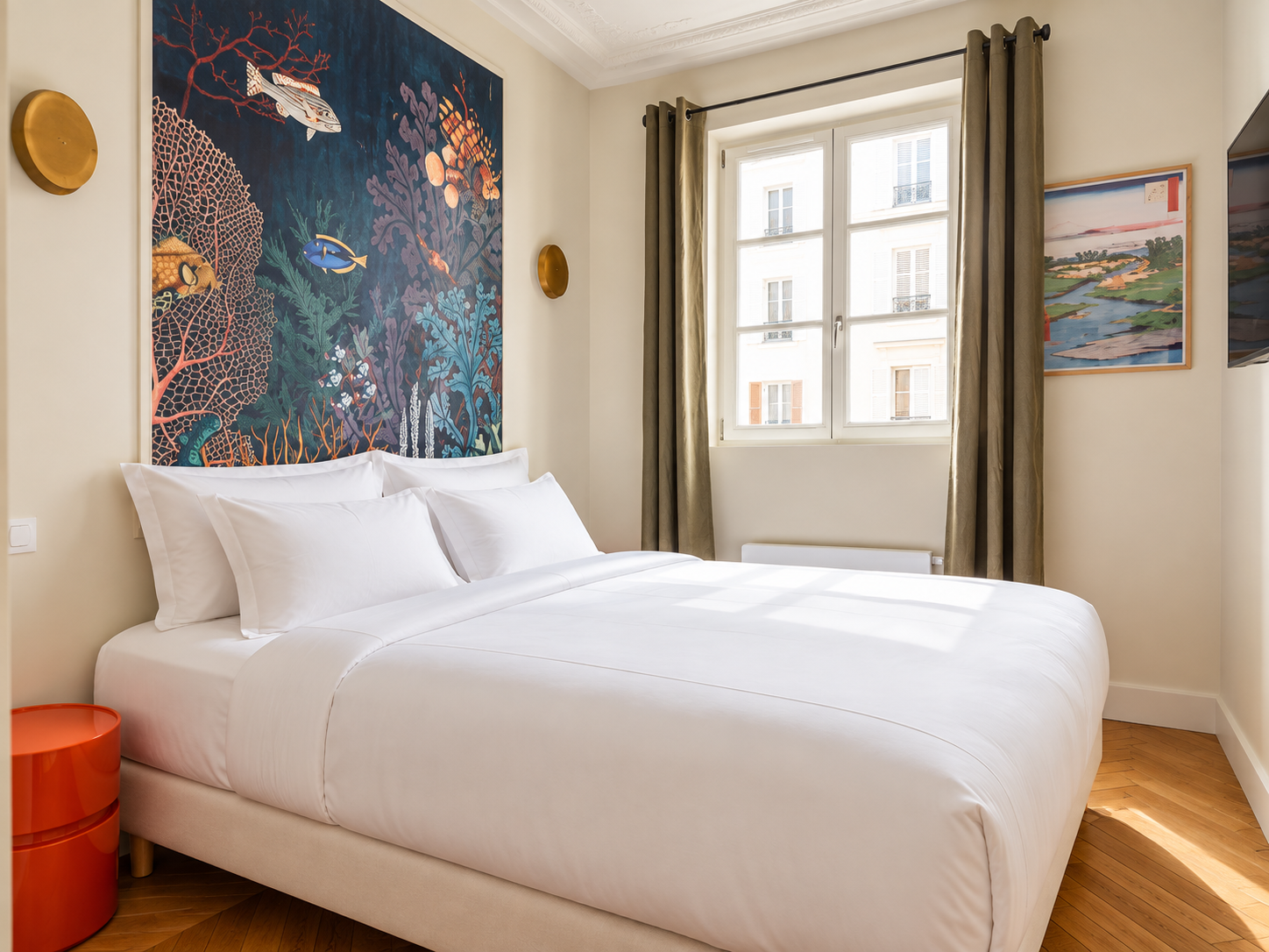 A bright bedroom with a white bed featuring white pillows and bedding, a colorful underwater-themed tapestry above the bed, green curtains on a large window, and framed landscape artwork on the walls.