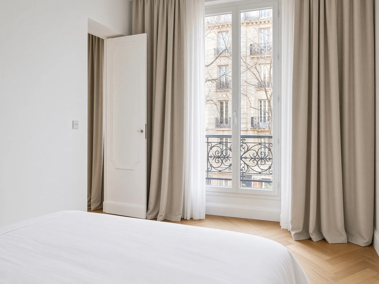 A bedroom with white walls, a balcony window with beige curtains, a white door, and a glimpse of the bed in the foreground.