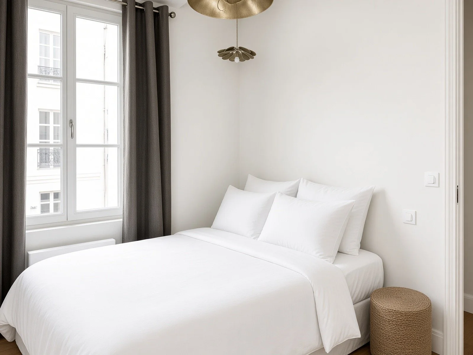 A bright, minimalist bedroom with white walls, a window with dark curtains, a neatly made bed with white bedding and pillows, and a wicker basket beside the bed.