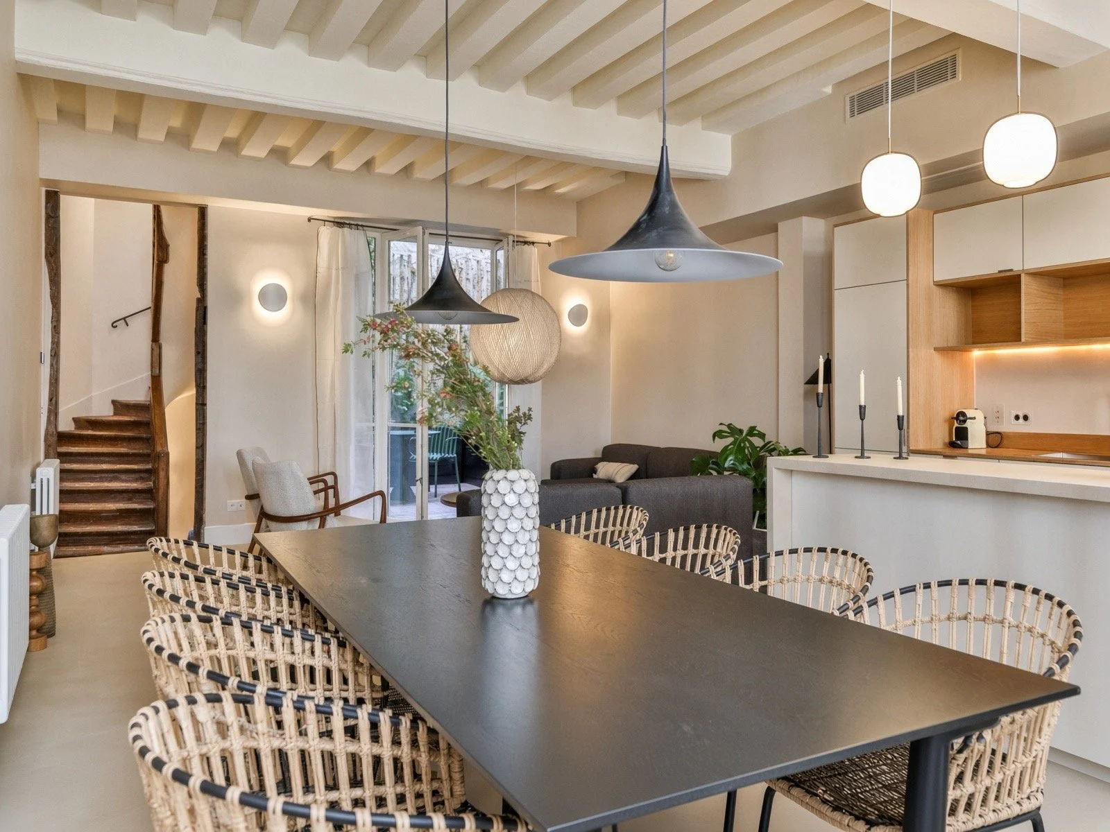 Modern open-concept living and dining area featuring a black dining table with woven rattan chairs, a gray sofa, pendant lights, and decorative accents including candle holders and a vase with flowers.
