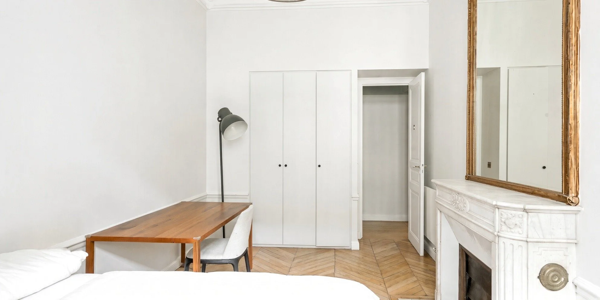 A corner of a bedroom with a wooden desk, a white chair, a black floor lamp, white walls, a white built-in closet, an open door, and a large gold-framed mirror above a decorative white fireplace.