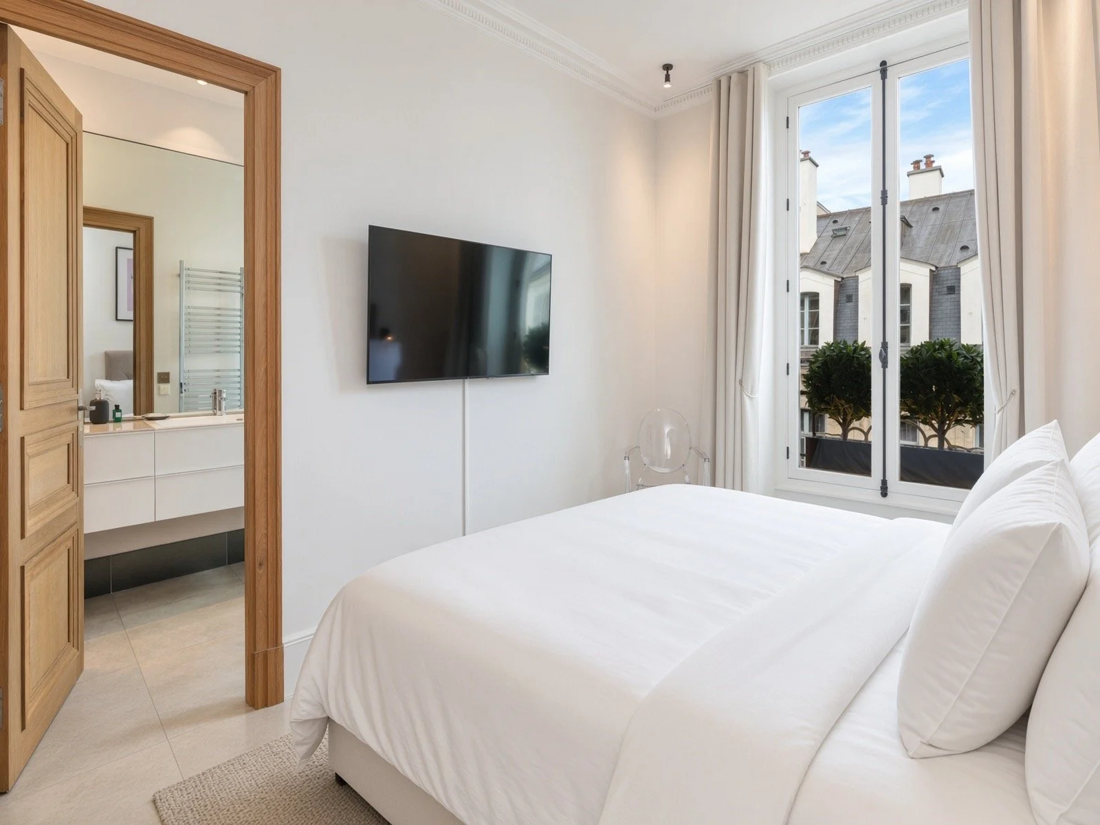 A bedroom with a large window, white walls, and a bed with white bedding. There's a flat-screen TV on the wall and a view of rooftops and trees outside.