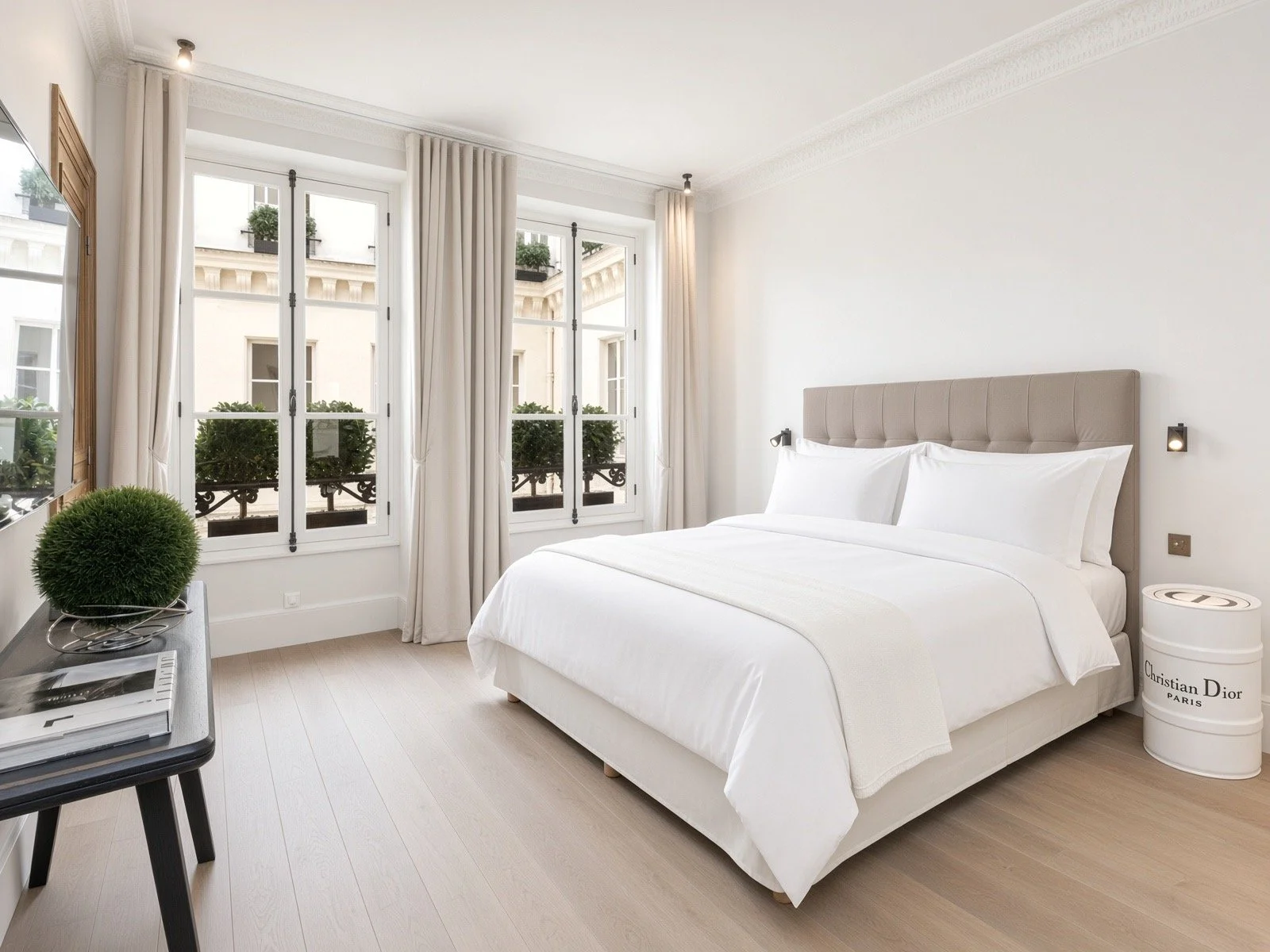 A bright bedroom with white walls, a large bed with white linens, and two windows with white curtains, overlooking a building with decorative architectural details. There is a small black table with green plant and magazines, black wall-mounted reading lights, and a round white side table labeled Christian Dior Paris.