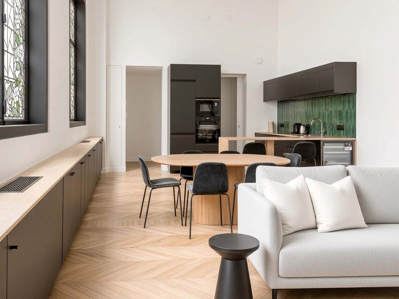 Modern kitchen and dining area with a round wooden table, black chairs, a white sofa with cushions, green tiled backsplash, black wall cabinets, and decorative stained glass windows.