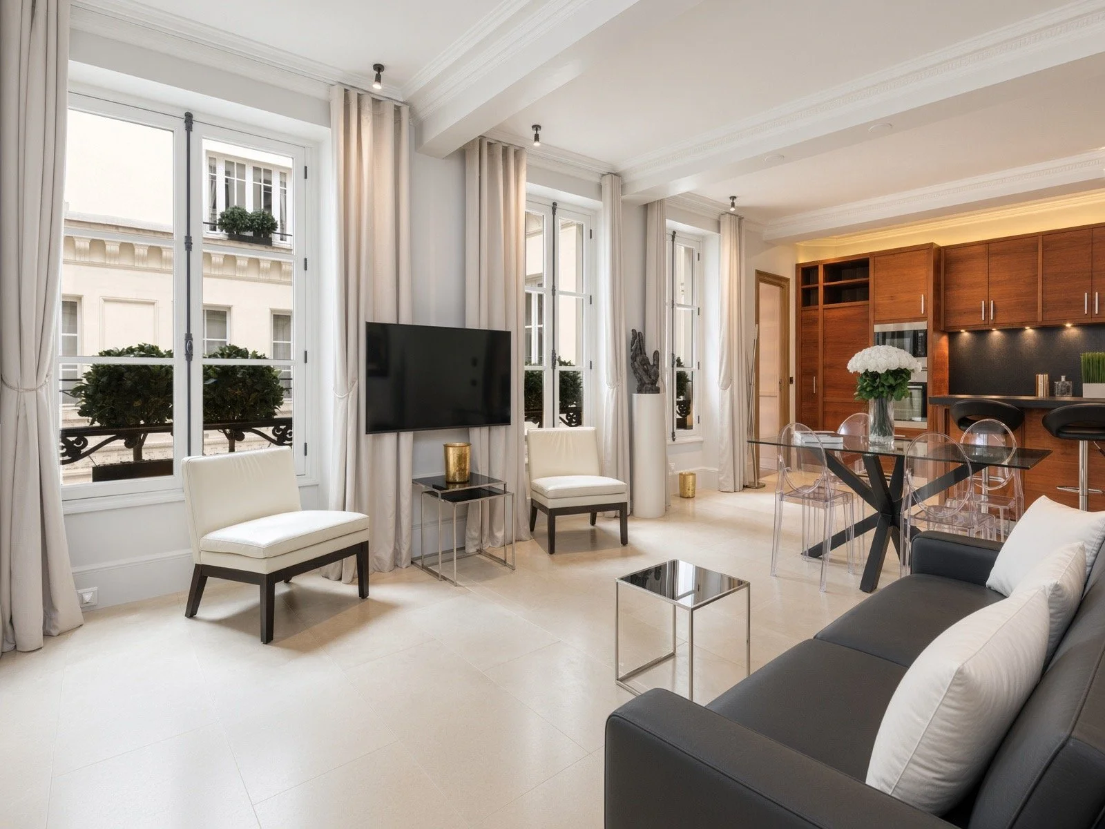 Modern living room with large windows, white curtains, white walls, a black and white sofa, glass coffee table, white chairs, a flat-screen TV, potted plants, and a wooden kitchen cabinet in the background.