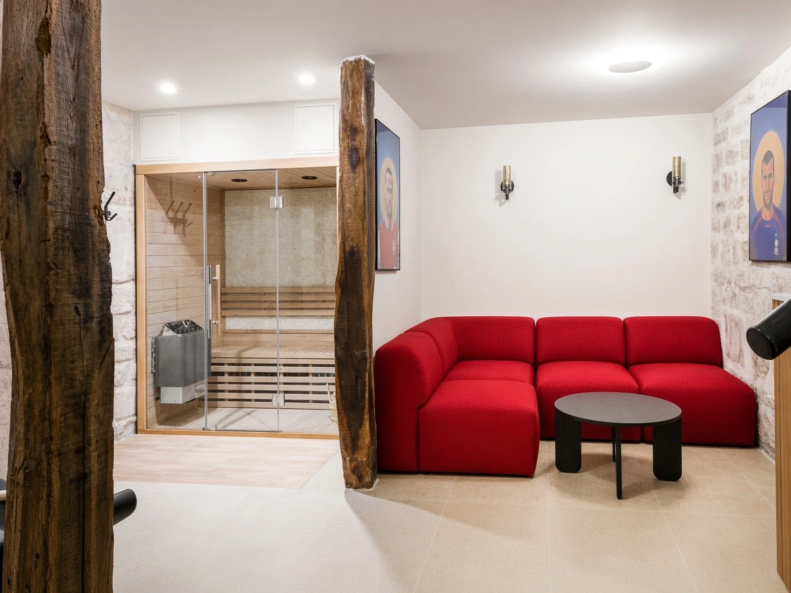 A cozy indoor lounge area with a red sectional sofa, a small dark coffee table, and a sauna room with glass doors, wood interior, and a built-in heater.