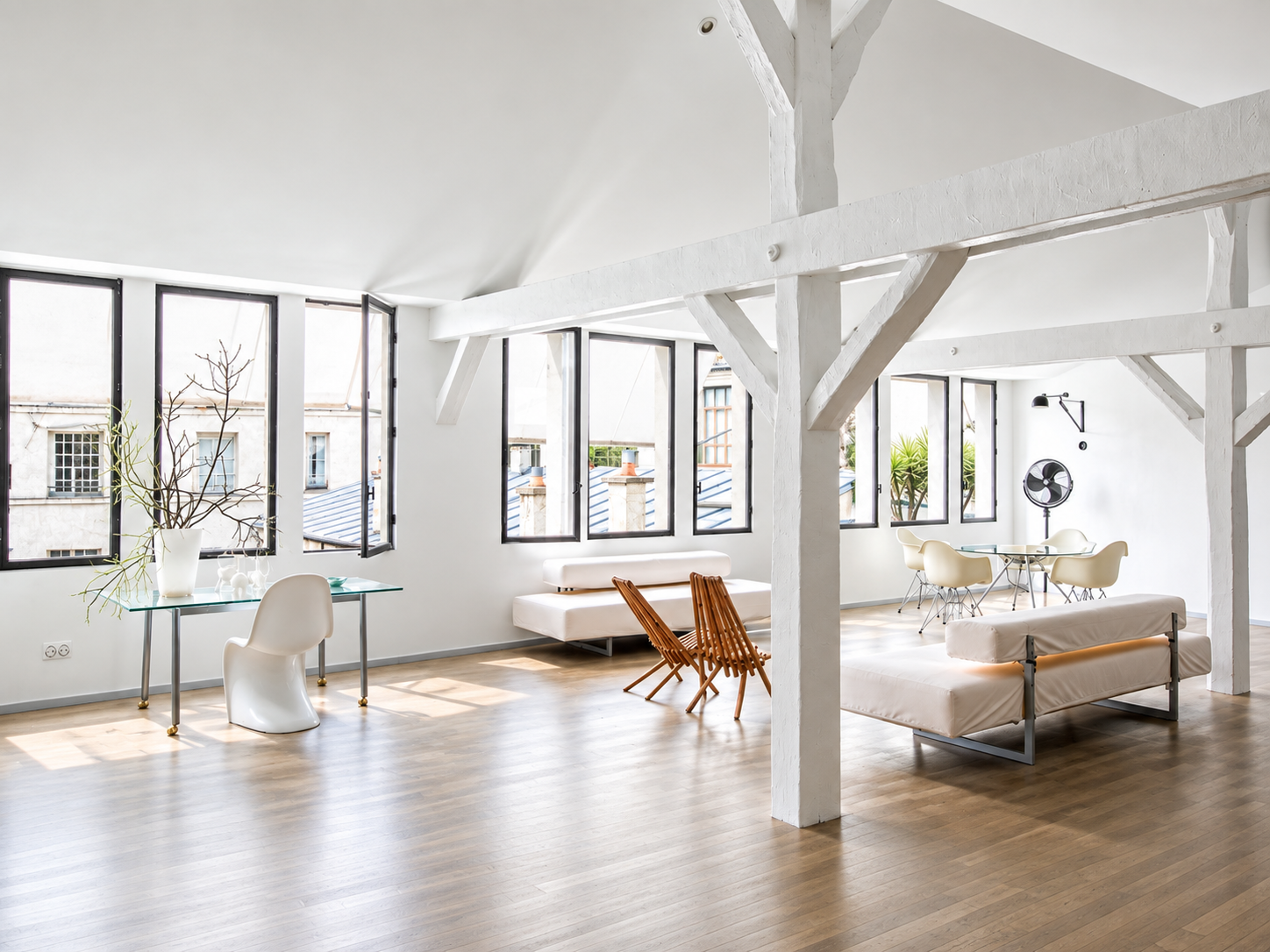 Bright, minimalist living space with white walls and large windows, showcasing a mix of modern and rustic furniture including a glass table with white chairs, wooden dining chairs, a beige sofa, and a white lounge chair, with a black fan and sunlight streaming in.