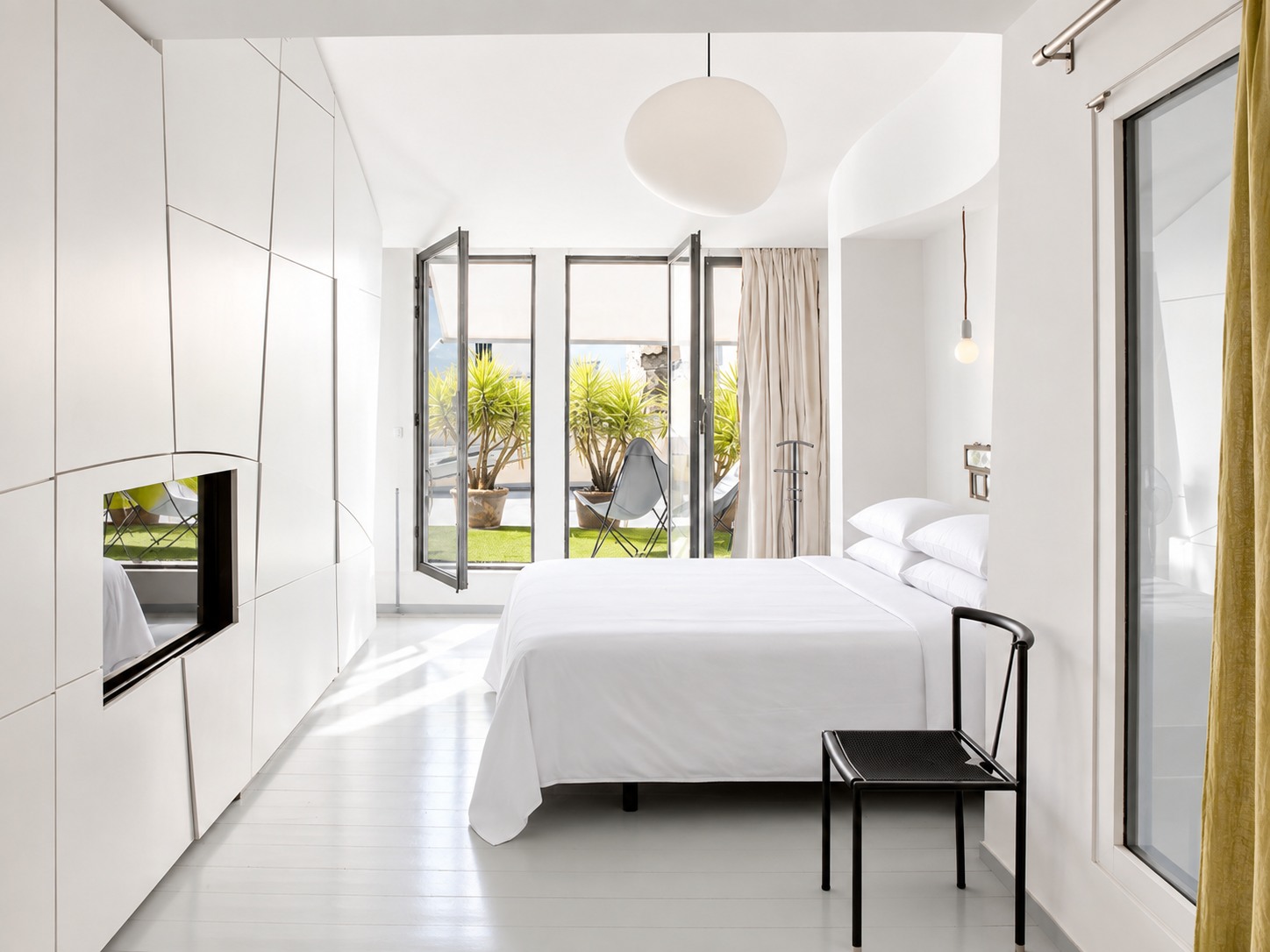 Modern white bedroom with a bed, black chair, and view of a small outdoor patio with potted plants