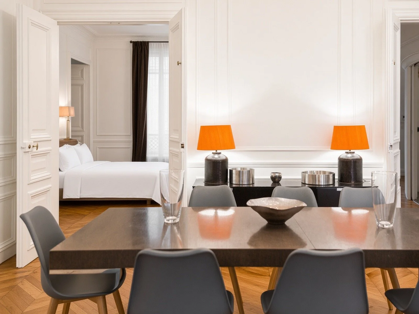Modern living and bedroom area with a dining table, six black chairs, two orange lamps on a black console, and a white bed with a nightstand and lamp in the background, with white walls and wooden flooring.