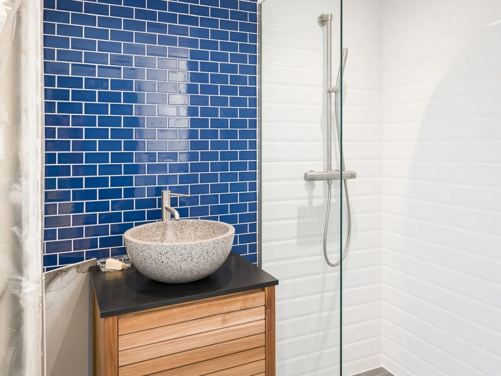 Modern bathroom with blue subway tile wall, white tile wall, concrete vessel sink on wooden vanity, and glass-enclosed shower with handheld showerhead.