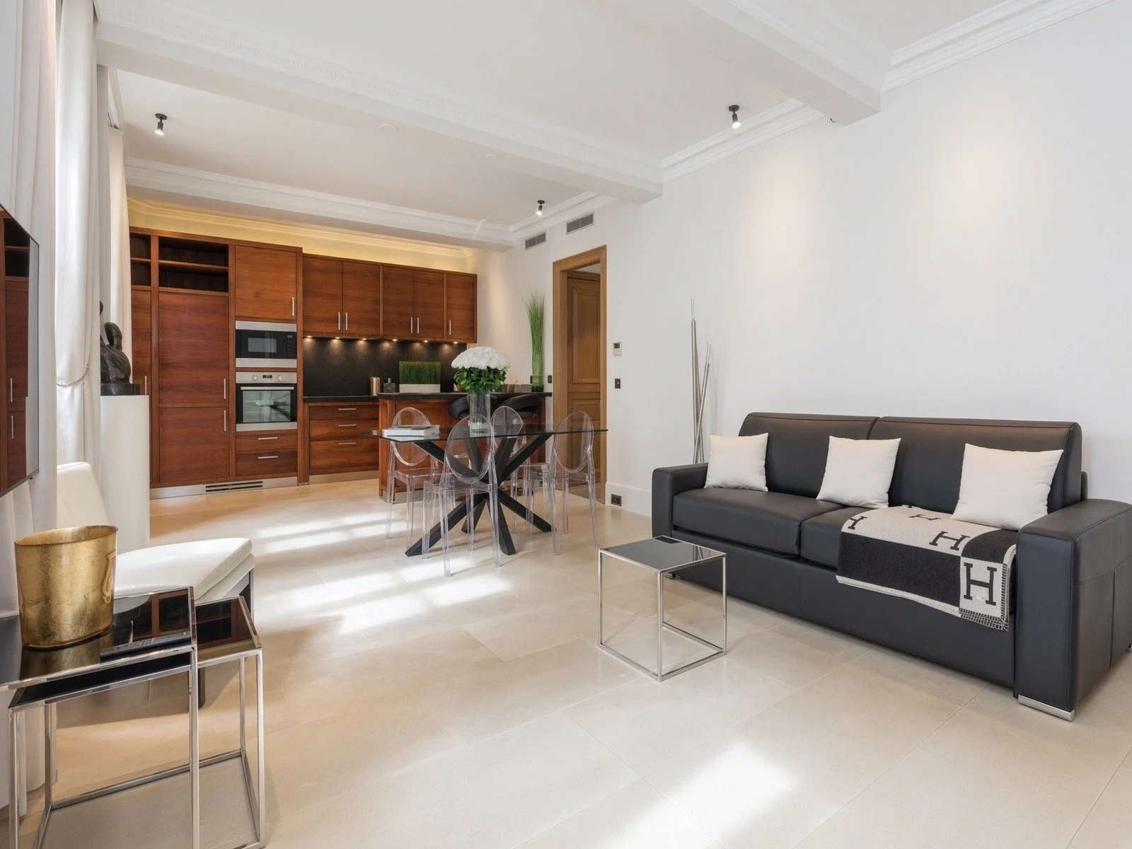 Modern living room with black sofa, white pillows, glass dining table with four clear chairs, and wooden kitchen cabinets in the background.
