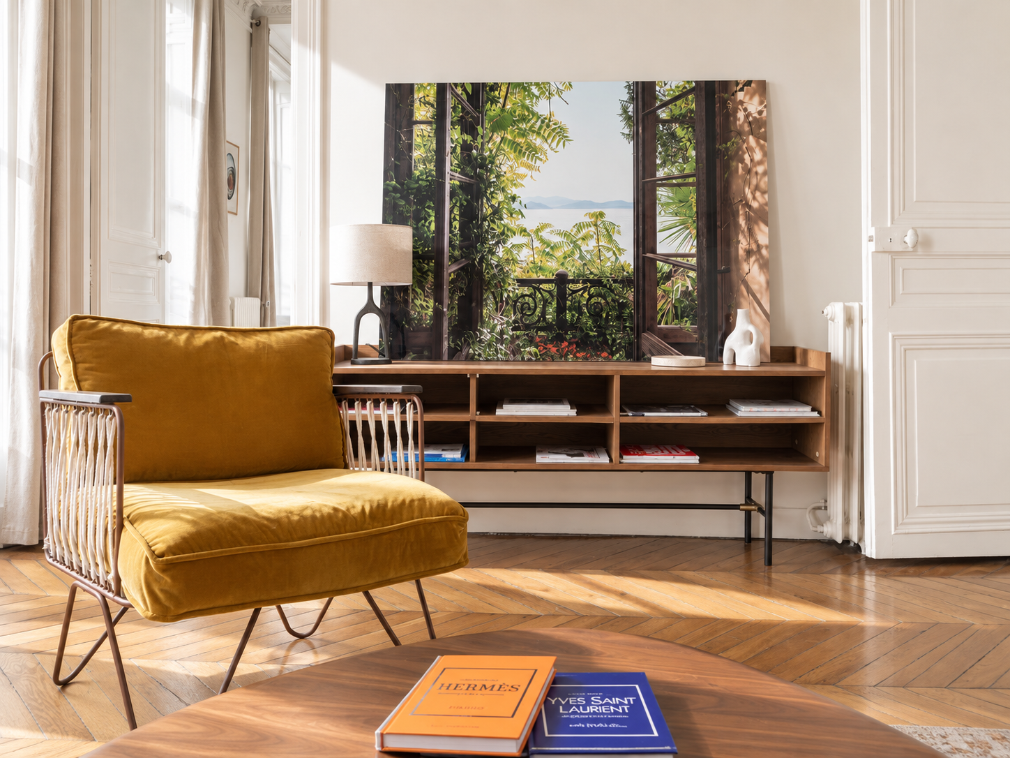 Living room with a yellow velvet armchair, a wooden TV stand with magazines, a table with Hermes and Yves Saint Laurent books, a table lamp, white decorative sculpture, and a large landscape painting of a balcony overlooking greenery and water.