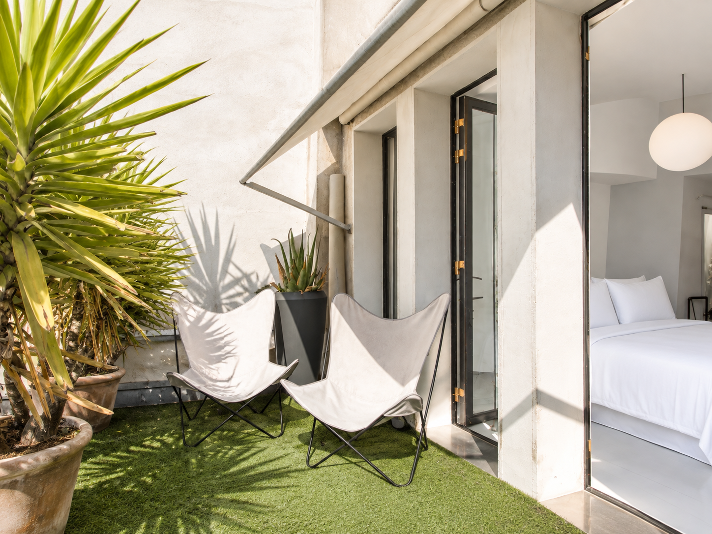 A small outdoor patio with two white butterfly chairs, potted plants, and an artificial grass floor, adjacent to a modern bedroom with white sheets and a hanging globe lamp.