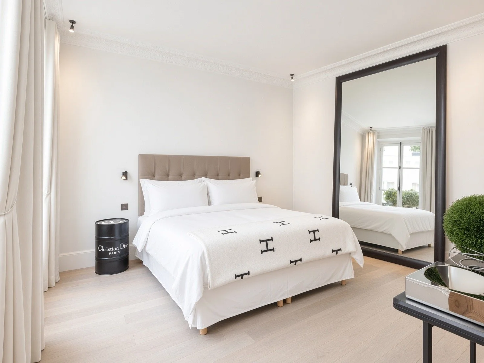 Modern bedroom with large mirror, white bedding with H pattern, beige headboard, black side table, green plant, large window with white curtains, and light wood flooring.