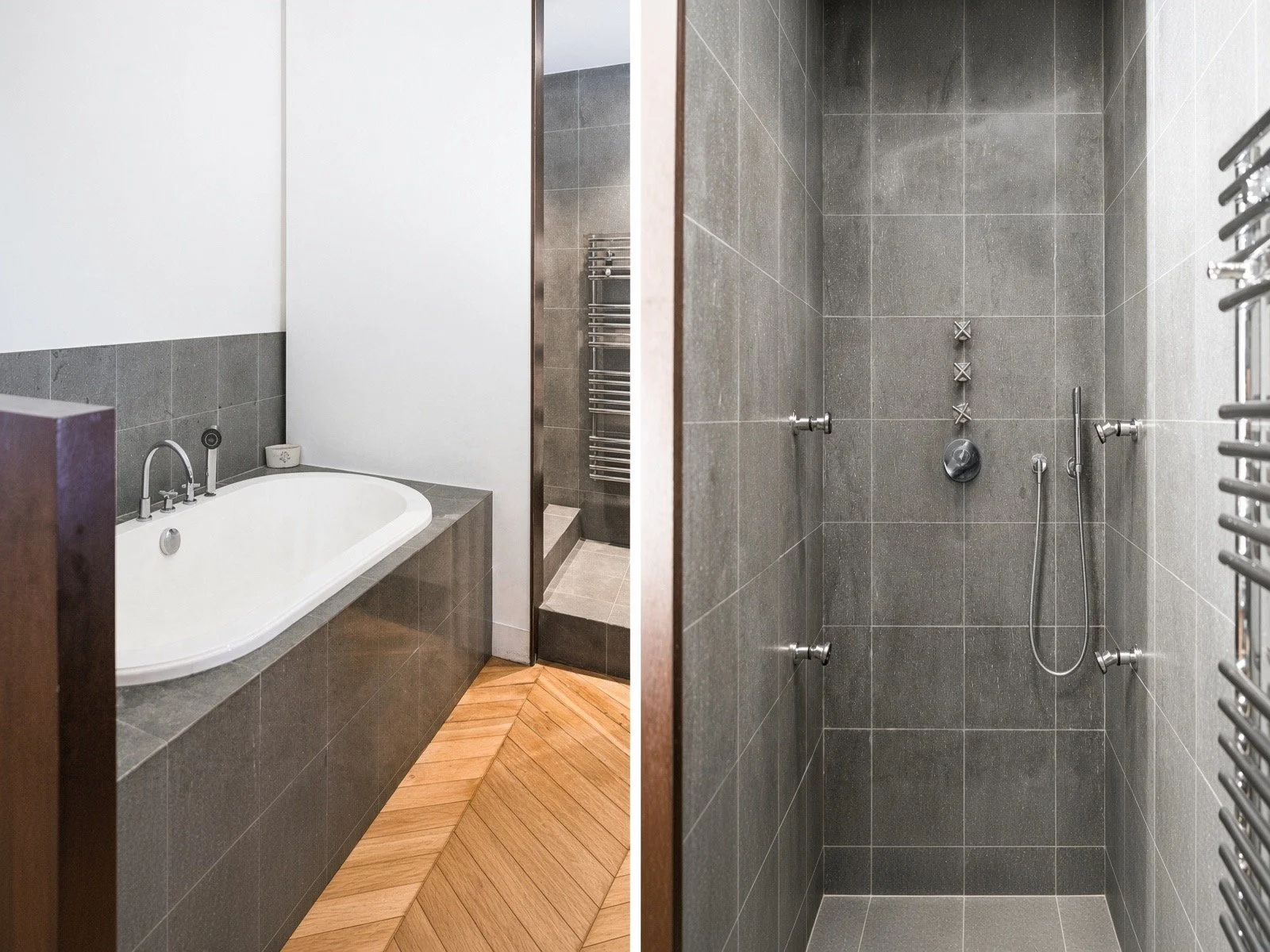 A bathroom with a built-in bathtub on the left side, surrounded by dark gray tiles and a wooden floor. On the right side, there is a shower area with dark gray tiled walls and a chrome shower fixture, including a handheld showerhead and multiple body jets.