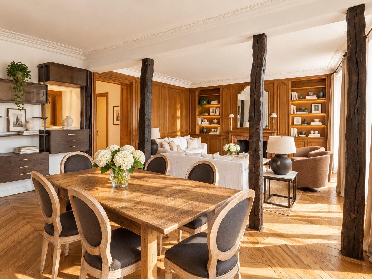 Beautifully decorated living room and dining area with wooden accents, a large wooden dining table with six chairs, a white sofa, armchairs, bookshelves with decorative items, and flowers on the table.