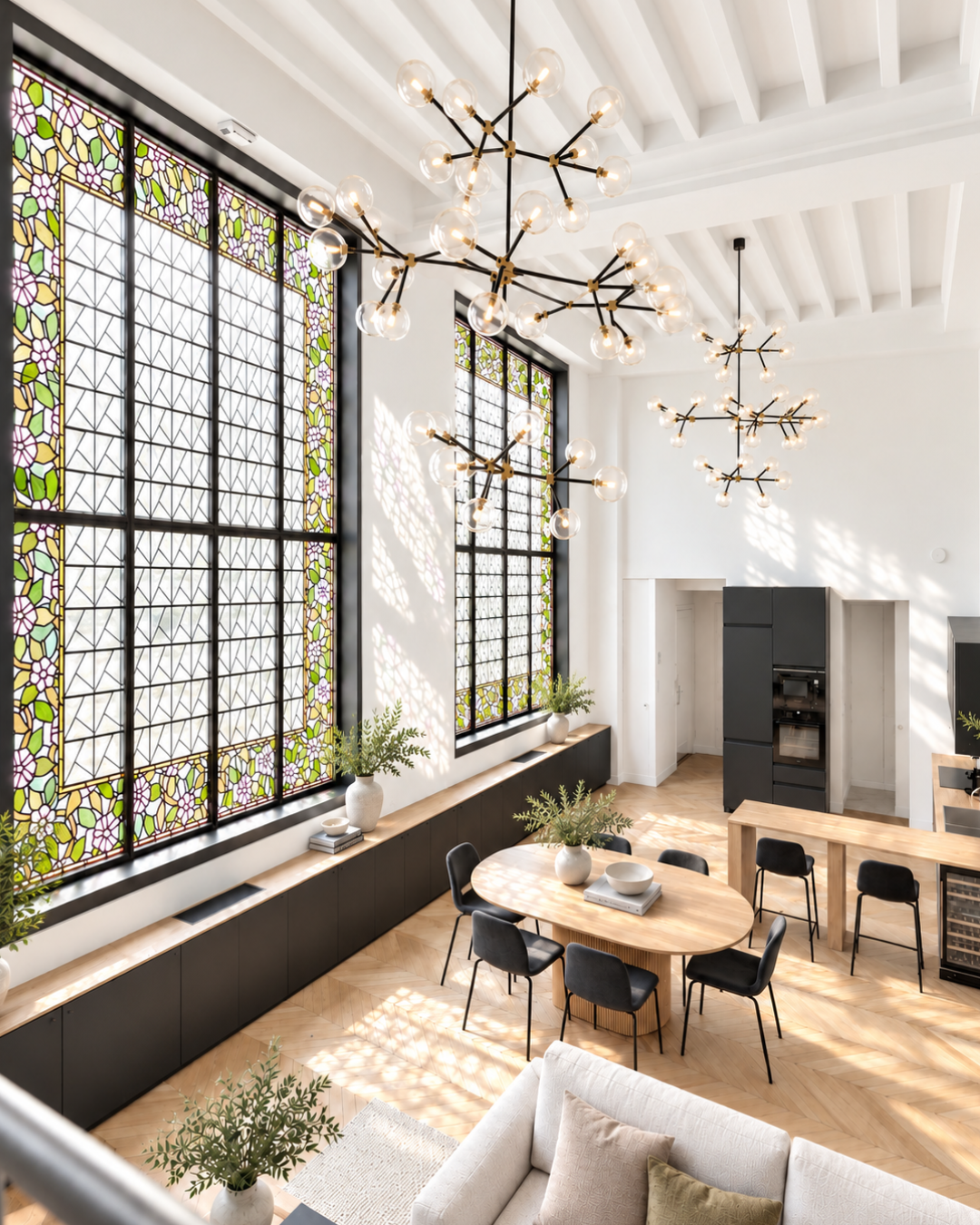 Bright living space with large stained glass windows, modern chandeliers, a dining table with black chairs, a beige sofa, and various plants.