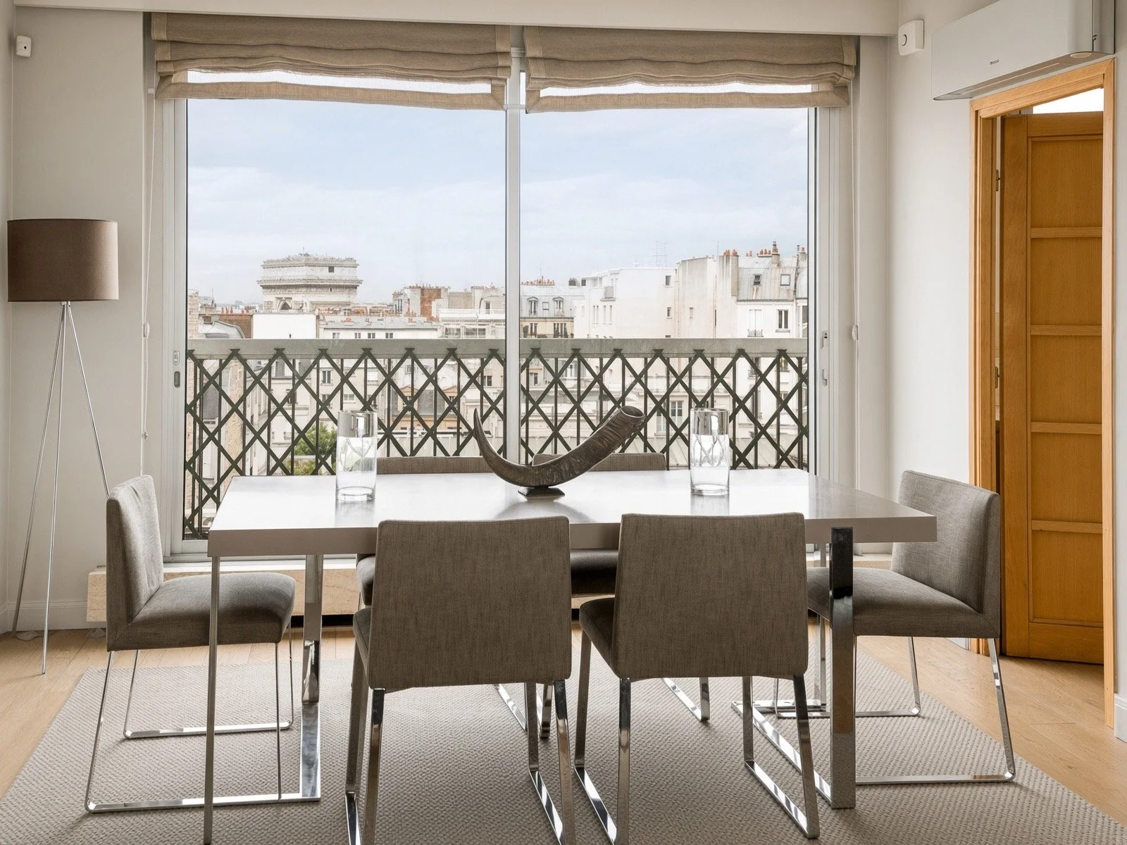 Dining room with a glass table, six gray chairs, a decorative centerpiece, and two glasses of water. Large sliding doors with a view of a city skyline and a black lattice balcony railing. A wooden cabinet on the right, a beige rug, and a floor lamp on the left.