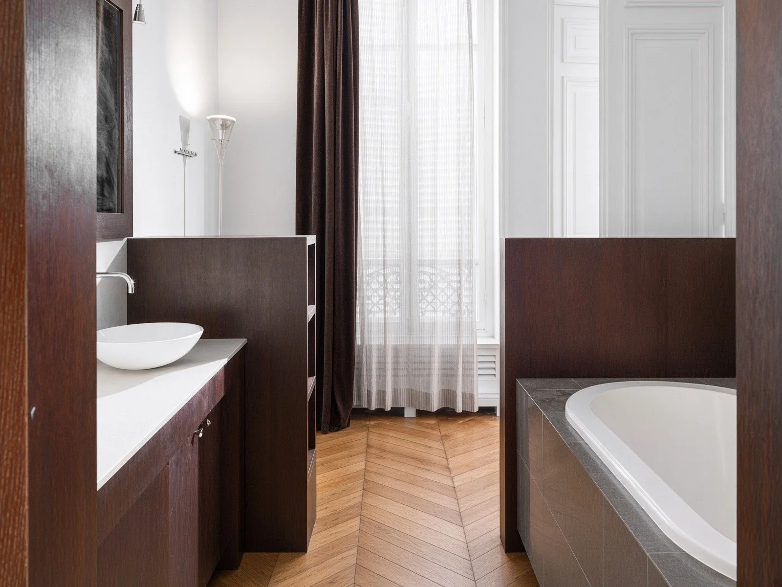 Modern bathroom with a round white sink on dark wood vanity, bathtub with gray tiled surround, tall window with sheer and dark curtains, wooden flooring, white decorative wall panels.
