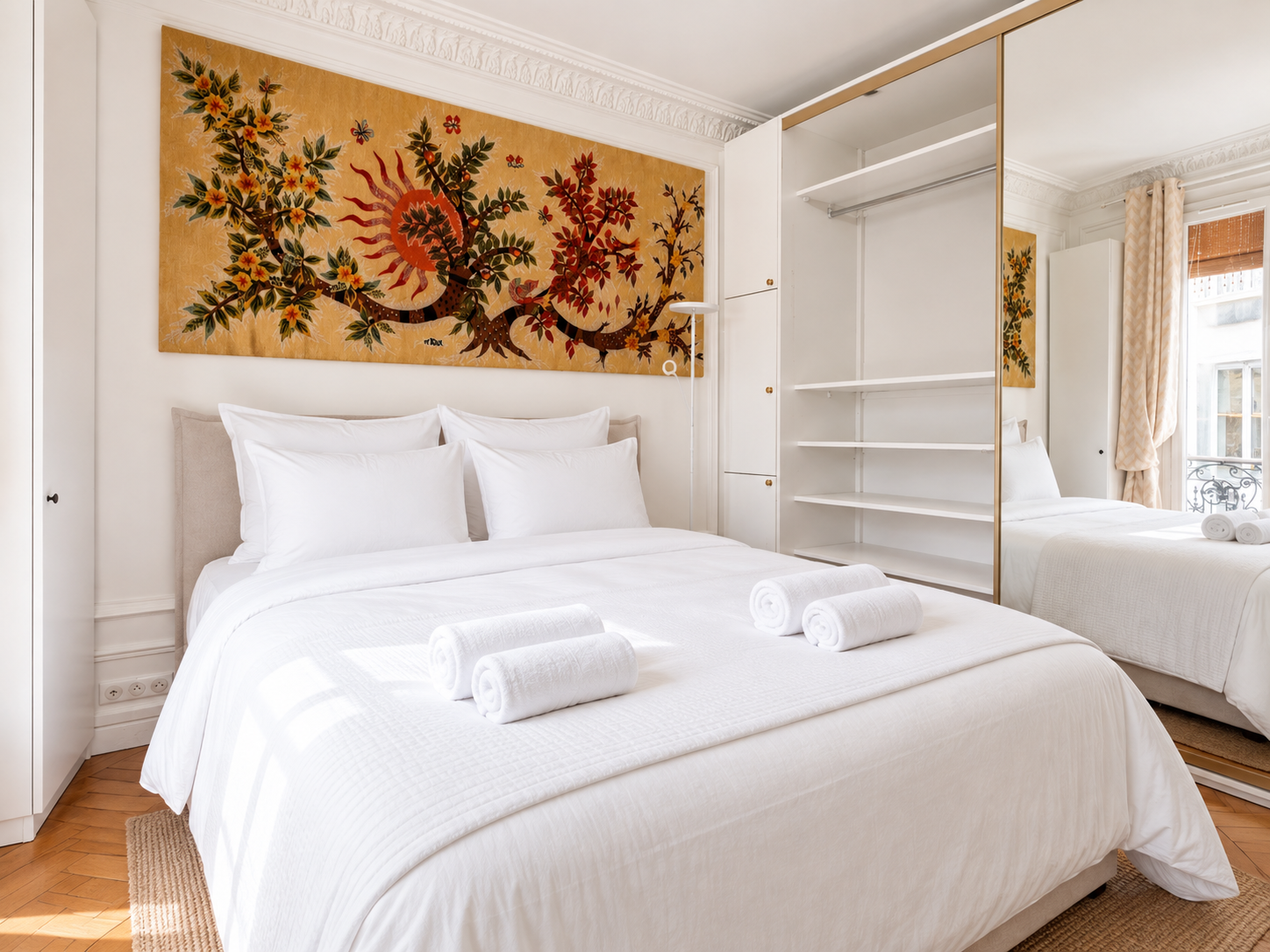 A bedroom with a large bed covered in white linens and pillows, three rolled white towels on top, a colorful wall art mural above, and an open closet with mirrored sliding doors reflecting a window with beige curtains.