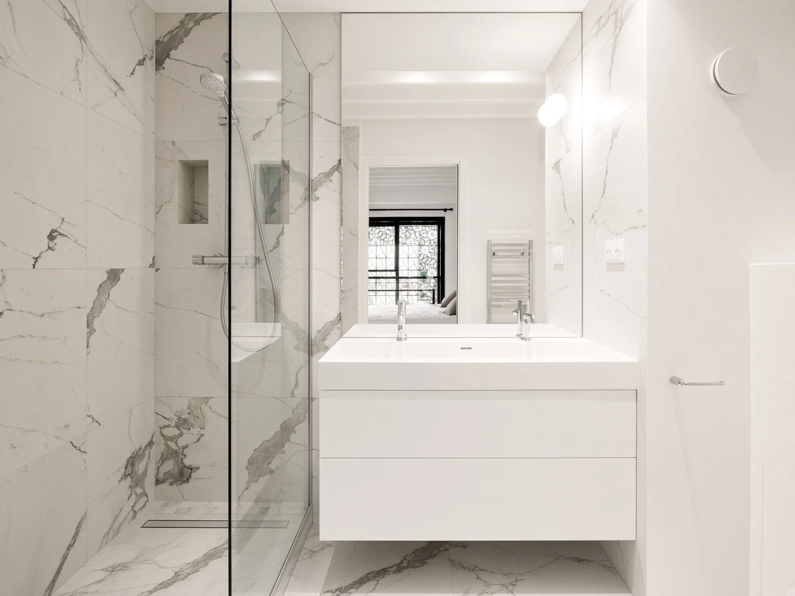 Modern bathroom with marble walls and floors, a walk-in glass shower with a handheld showerhead, a double sink vanity with a large mirror, and a window with a view of an outdoor balcony.