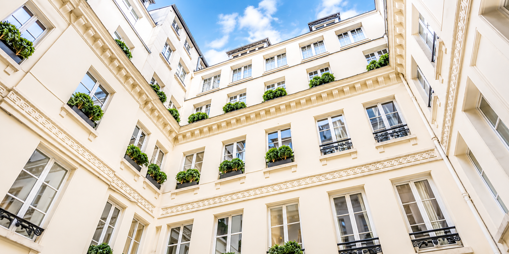 Photo of a courtyard in a multi-story beige building with large windows and green plants in window boxes, taken from the ground looking up towards a blue sky with clouds.