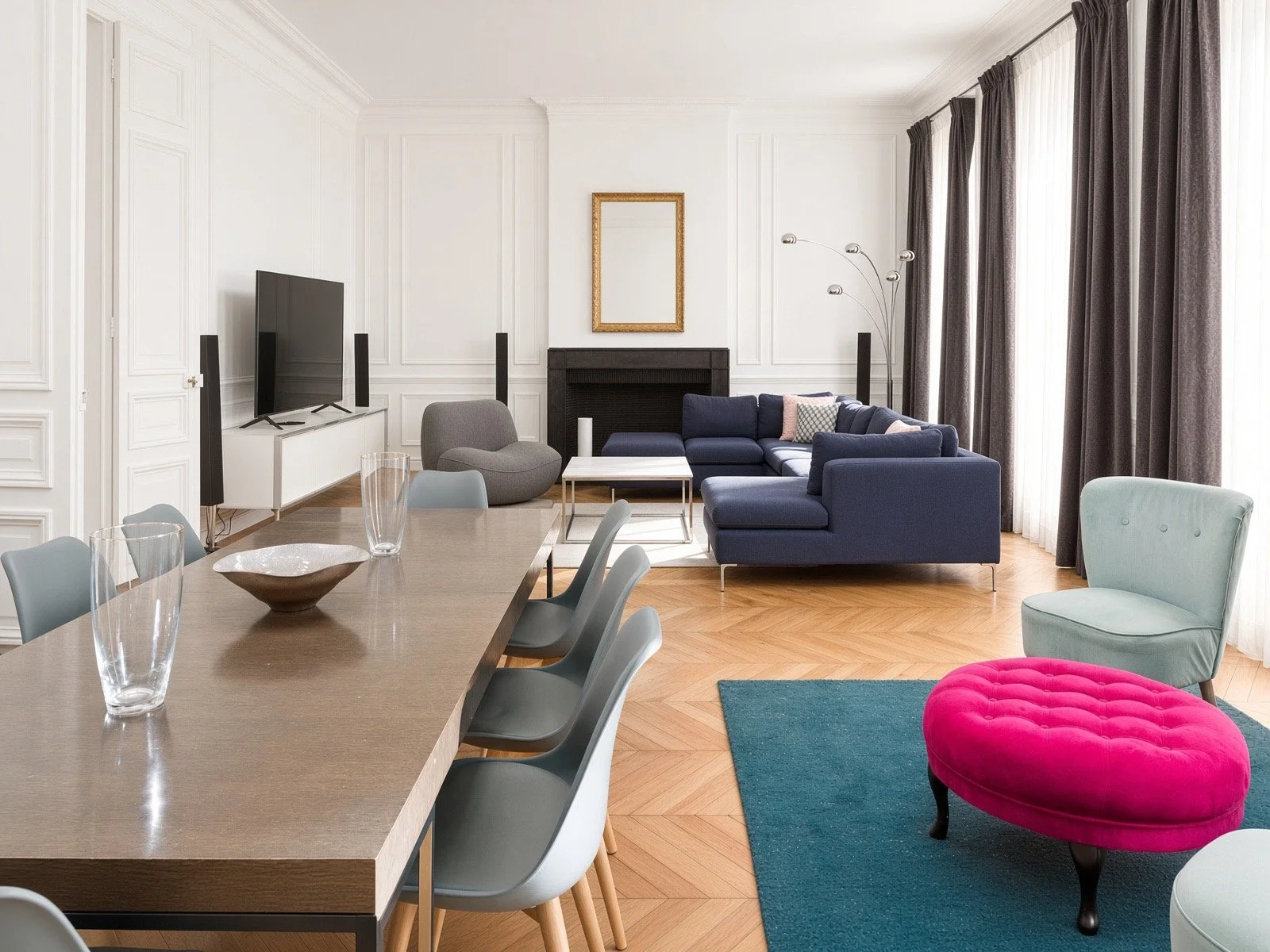 Living room with white paneled walls, a blue sectional sofa, a gray lounge chair, a creamy green chair, a pink ottoman, a wooden dining table with gray chairs, a television with speakers, a fireplace, a glass coffee table, and large windows with dark curtains.