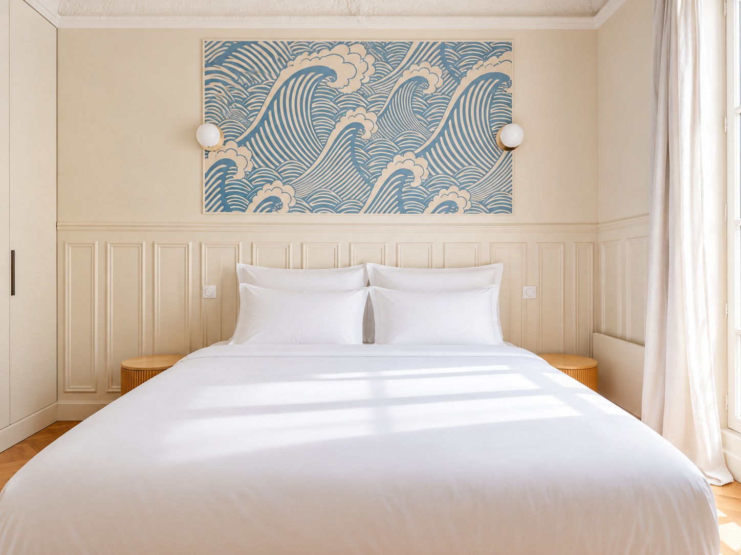 A neatly made bed with white bedding and pillows in a bedroom with cream-colored walls. There is a decorative blue and white wave art piece above the bed, two wall-mounted round lights on either side, and natural light coming through a window with white curtains.