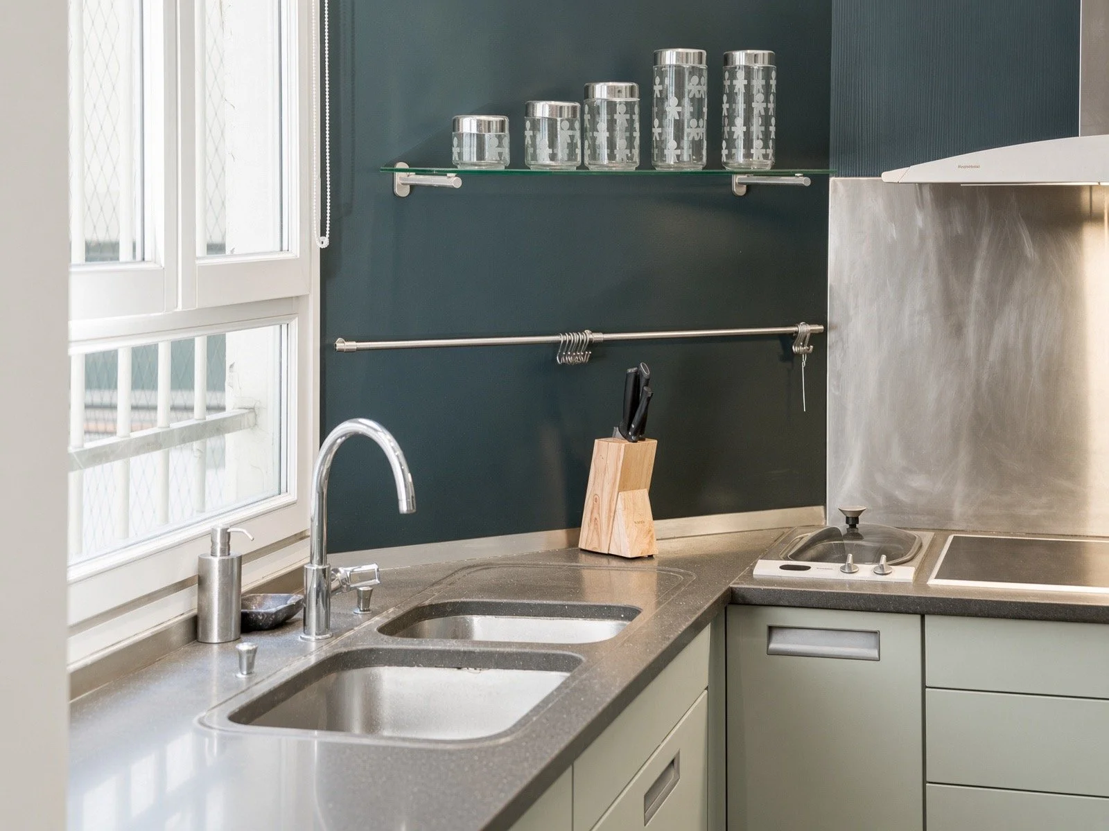 Kitchen with stainless steel sink, dark green cabinets, and a dark countertop. Glass jars with silver lids on a glass shelf above the counter. Knife in a wooden knife block, window with white frame, and a stove with a vent on the right.