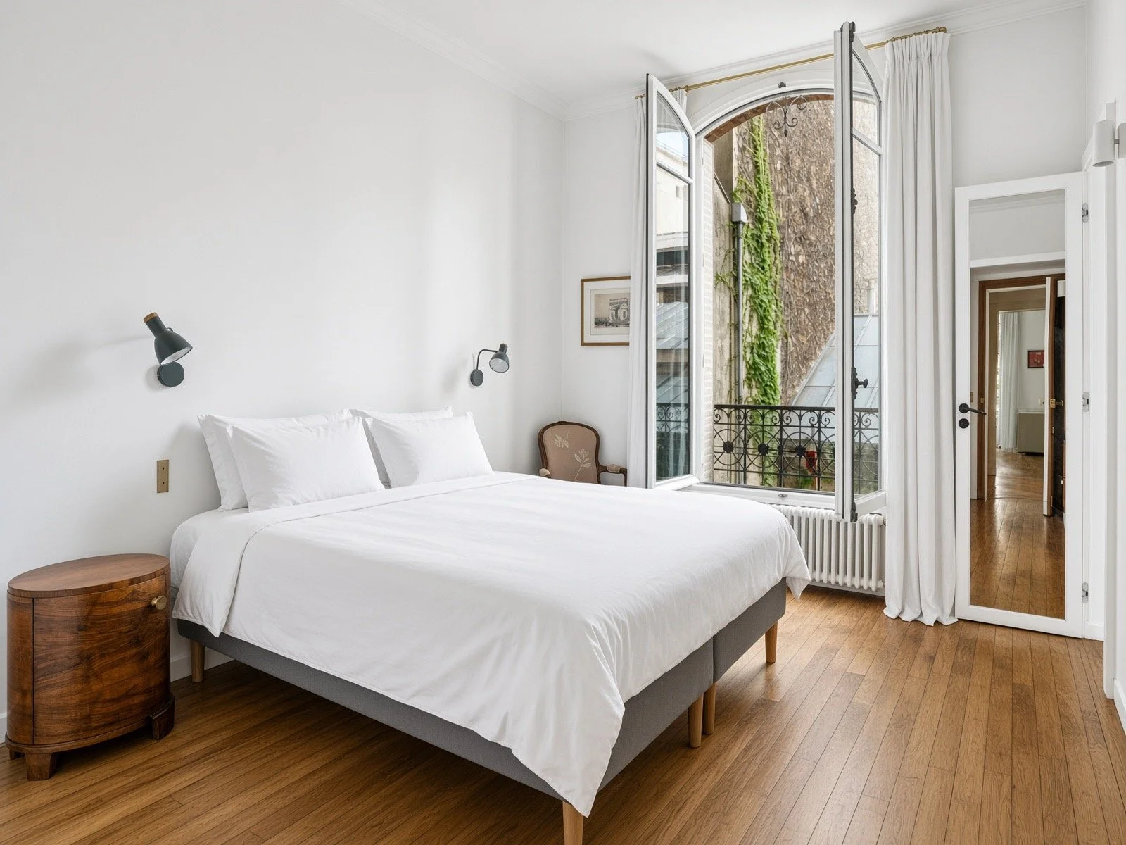 Bright bedroom with white walls, a large bed with white linens, a small wooden side table, and a vintage armchair. Large arched window with open white curtains, overlooking a balcony with greenery outside.