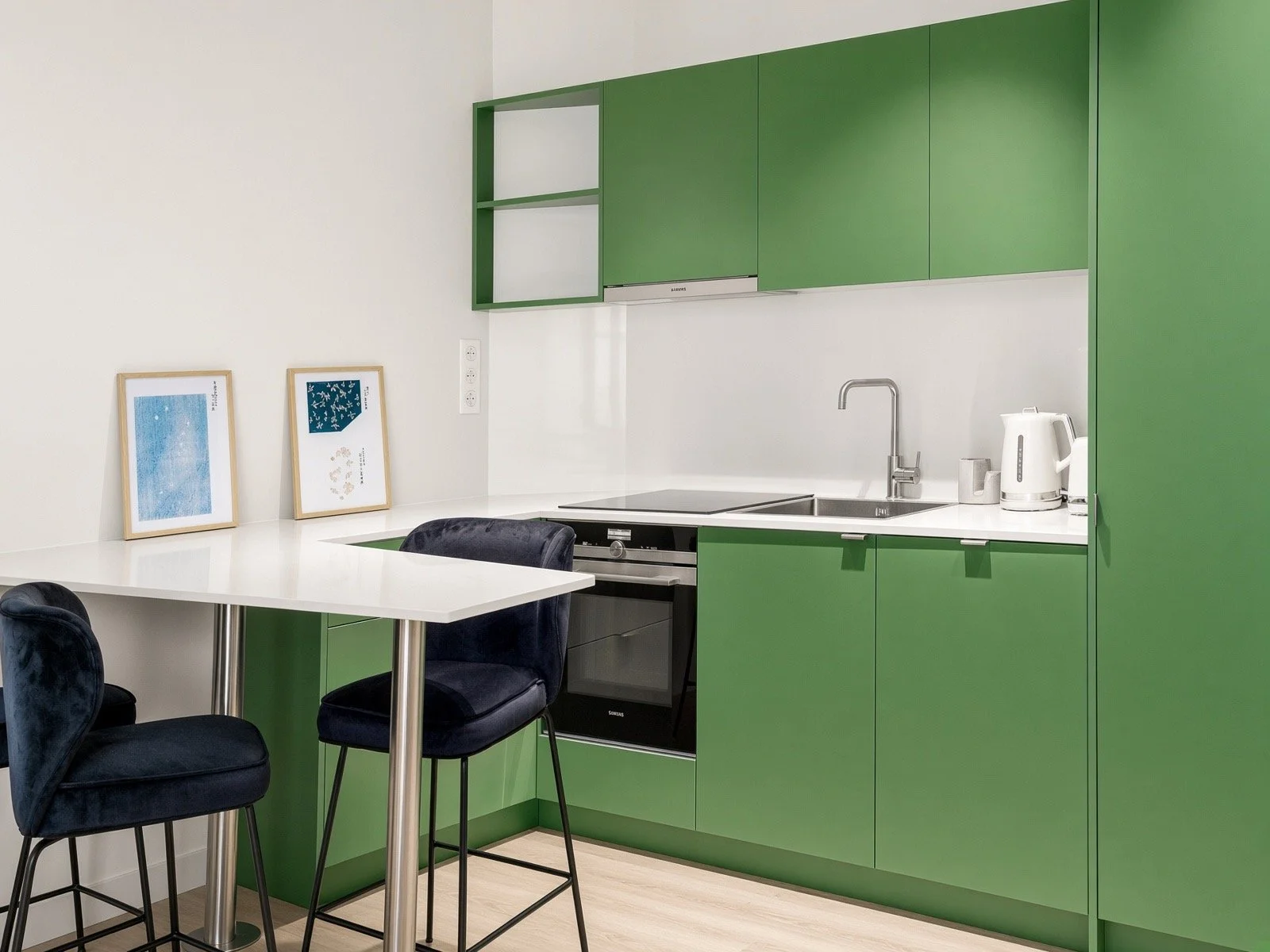 Modern kitchen with green cabinets, white countertop, black stove, and black chairs around a white dining table. Two framed artworks lean against the wall.